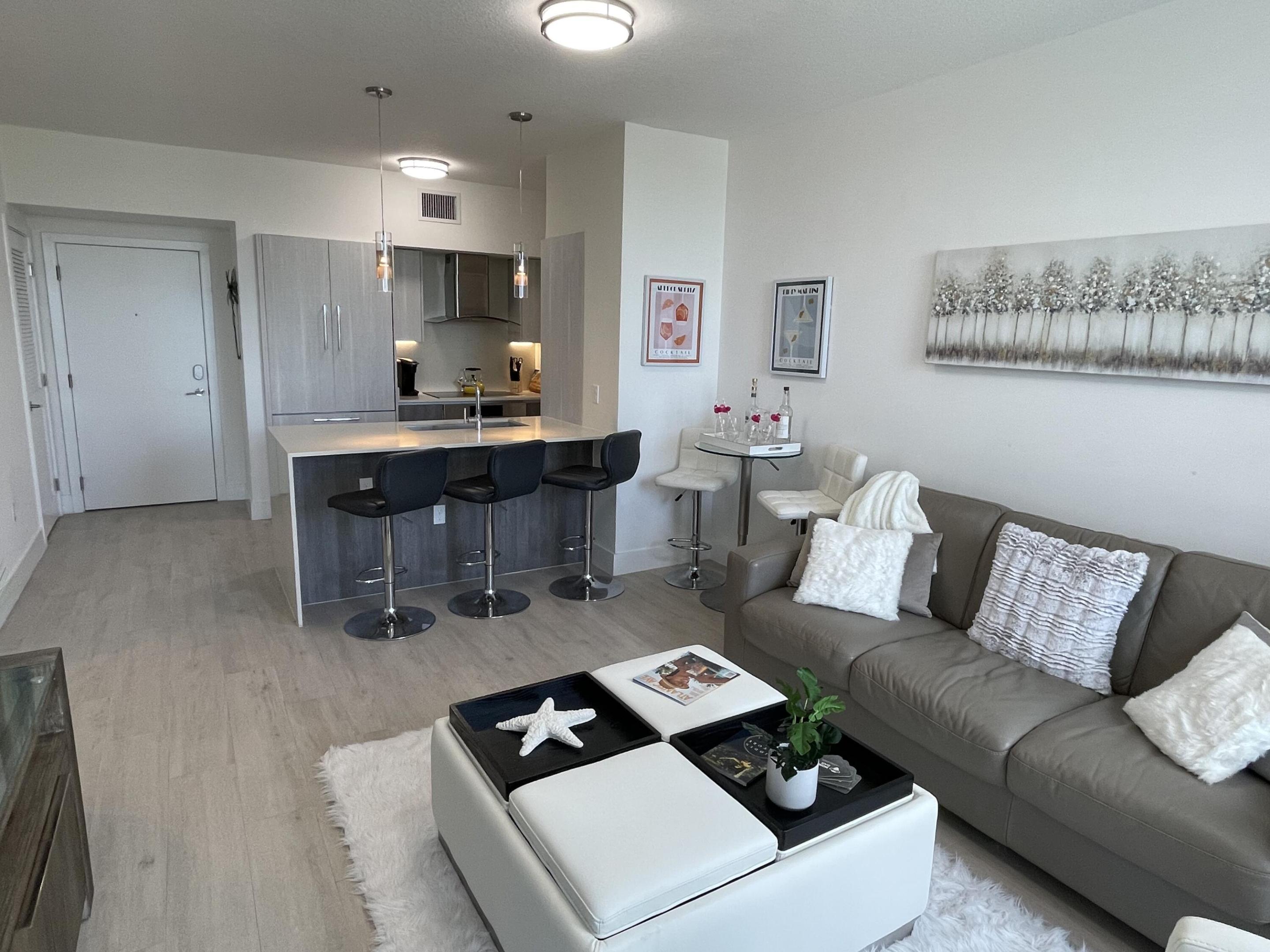 Open-plan living room and kitchen with a gray sofa, white coffee table, and island with three bar stools.