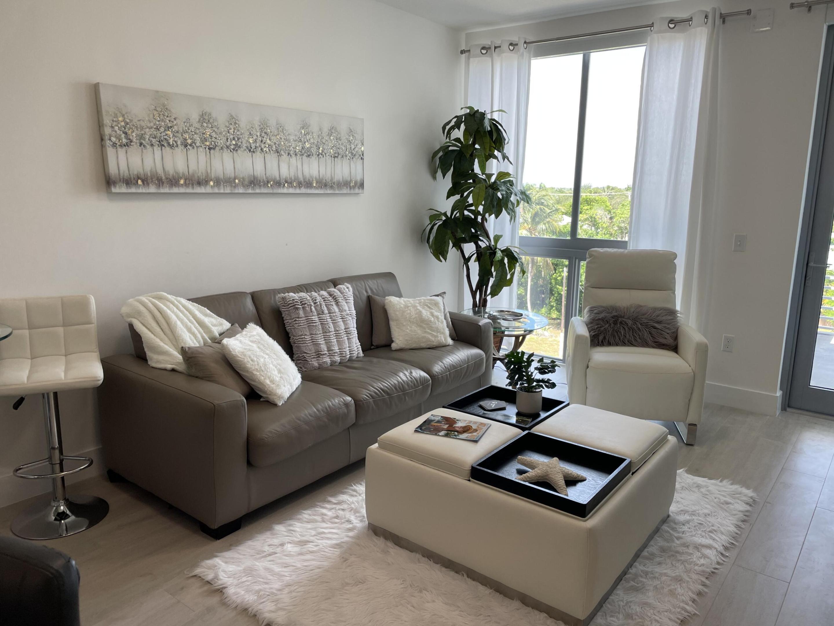 Staged living room with a gray sofa, white armchair, and large window overlooking trees.