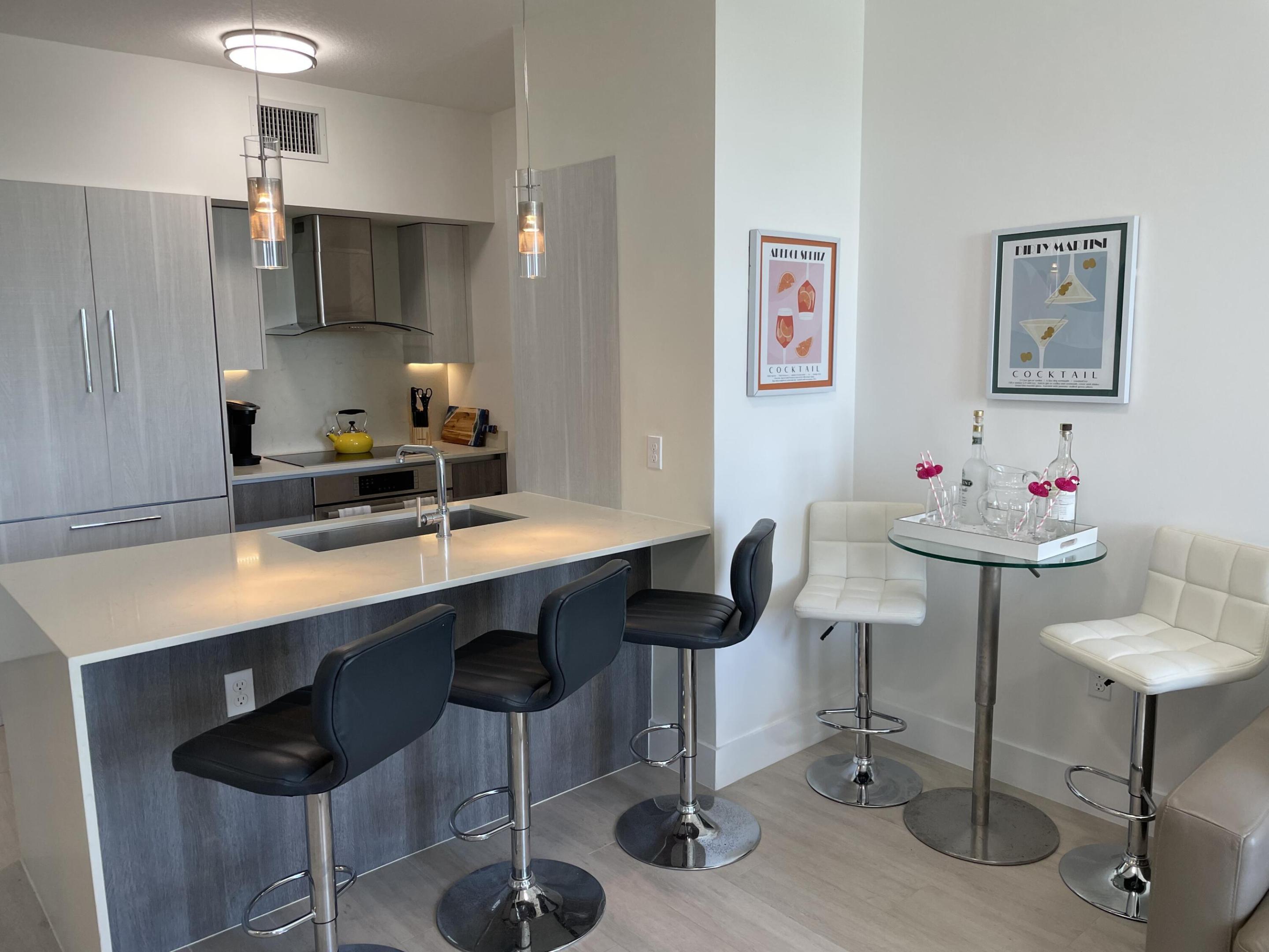 Modern condo kitchen with gray cabinets, a white breakfast bar, black stools, and a cocktail table with white chairs.