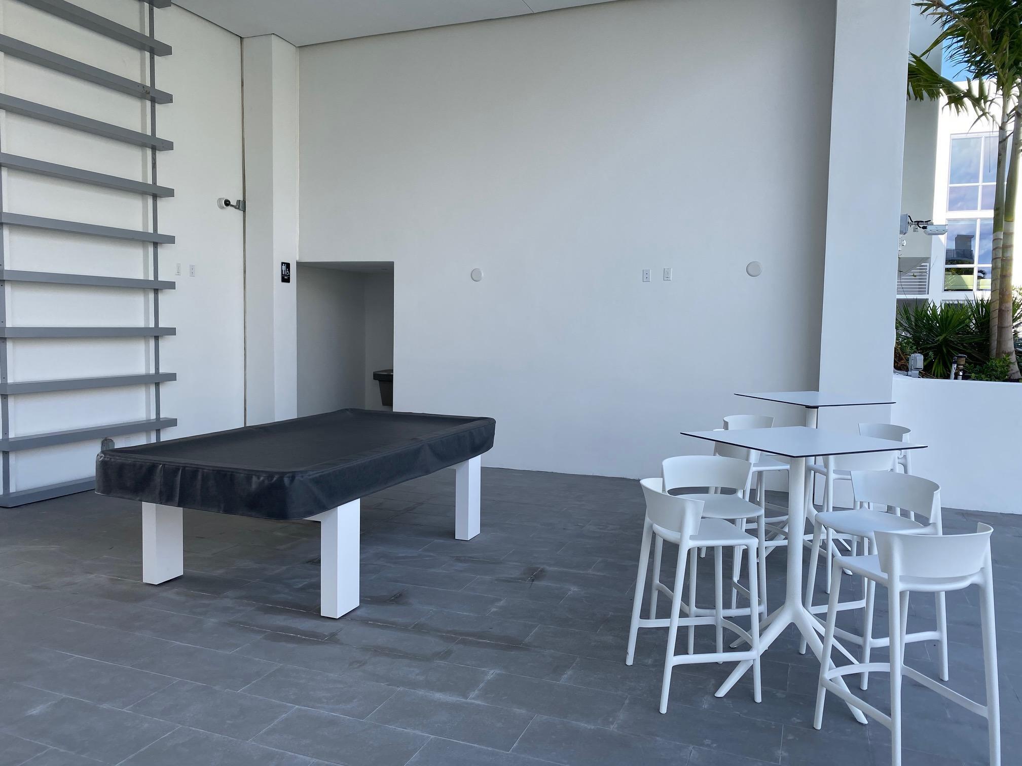 Outdoor amenity patio with a black pool table and white café seating beside tropical landscaping.