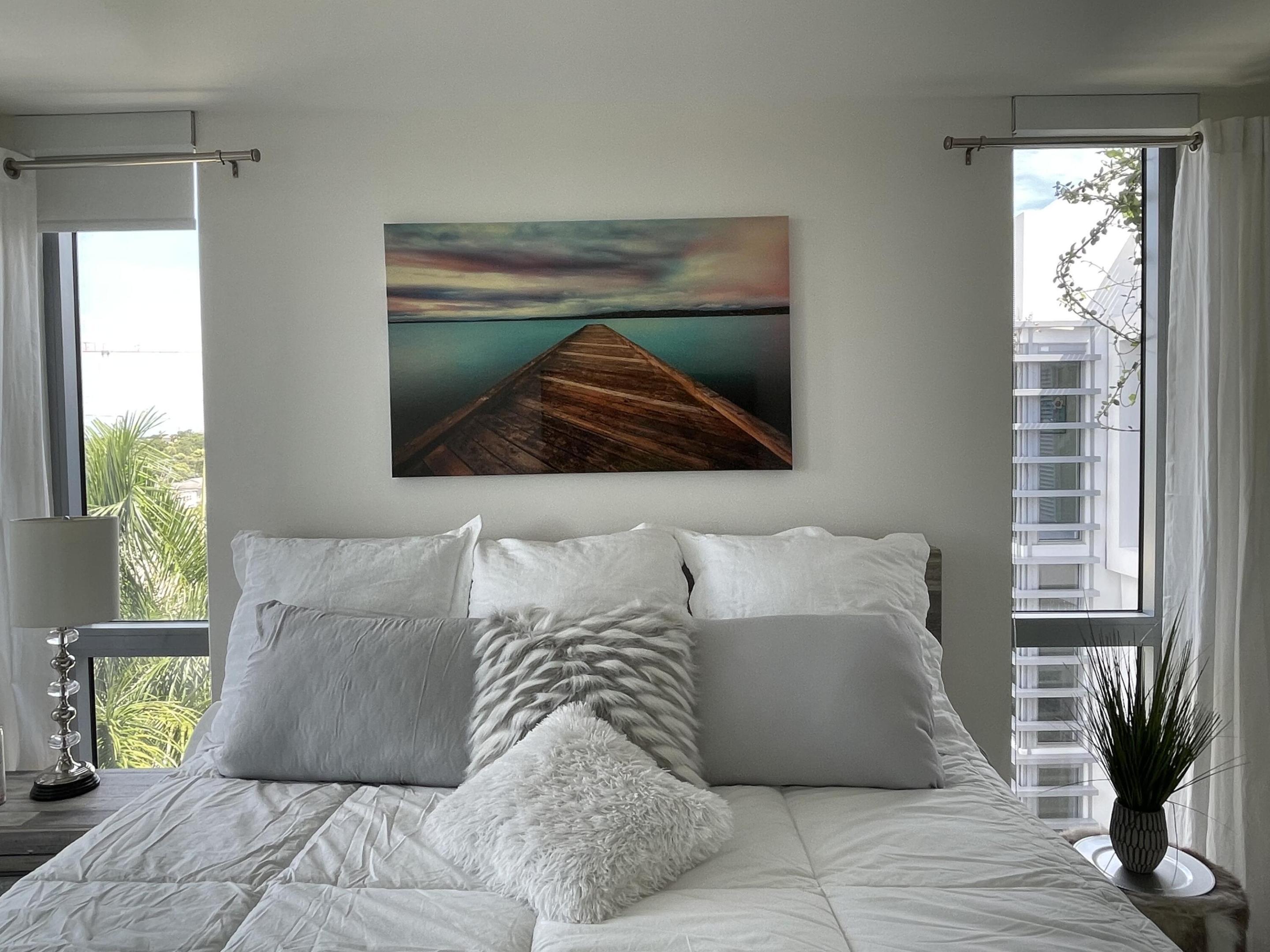 Bedroom with a white bed, pier artwork, and tall windows overlooking palms and a neighboring building.