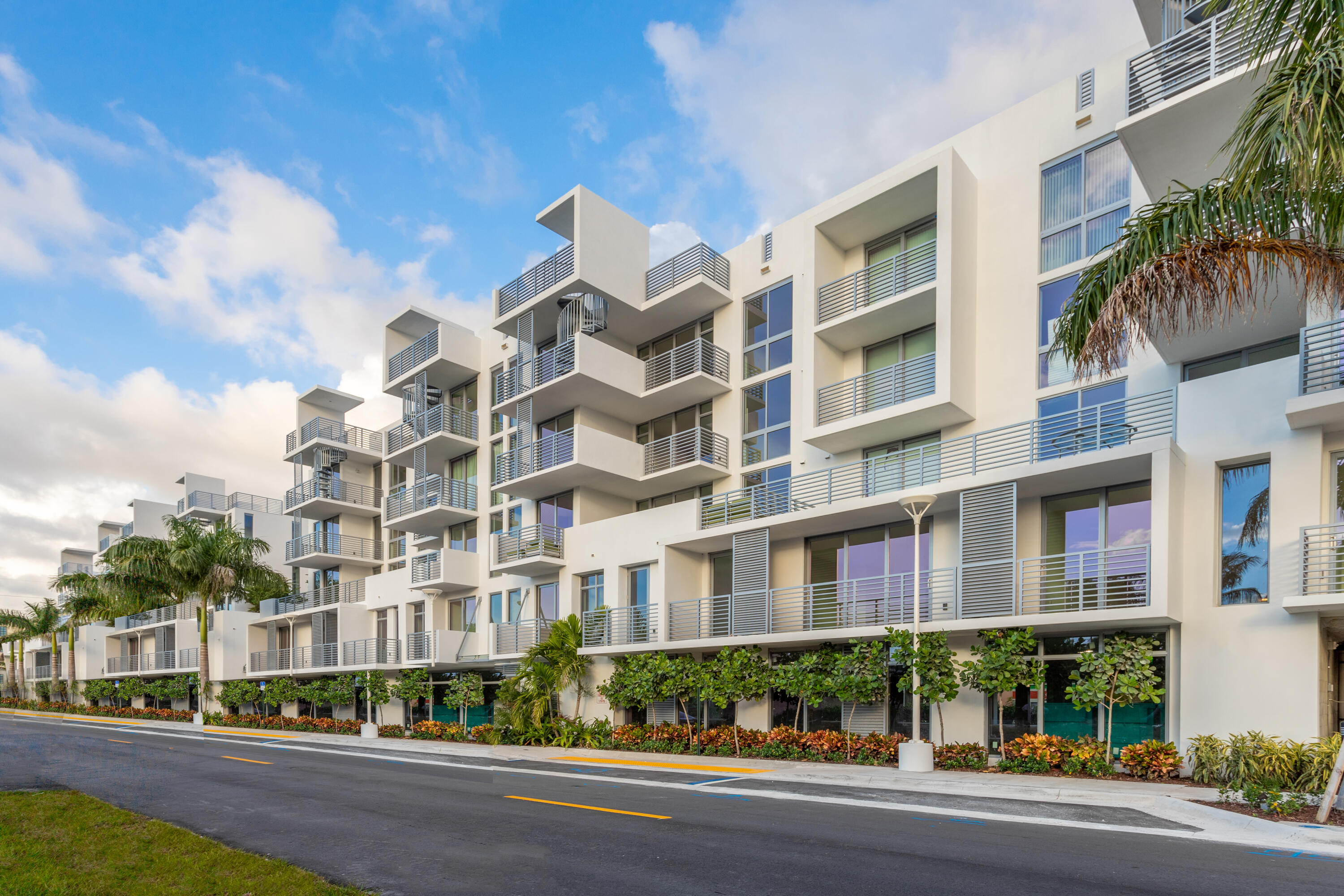 Modern white condominium building with stacked balconies, palm trees, and landscaped street frontage.