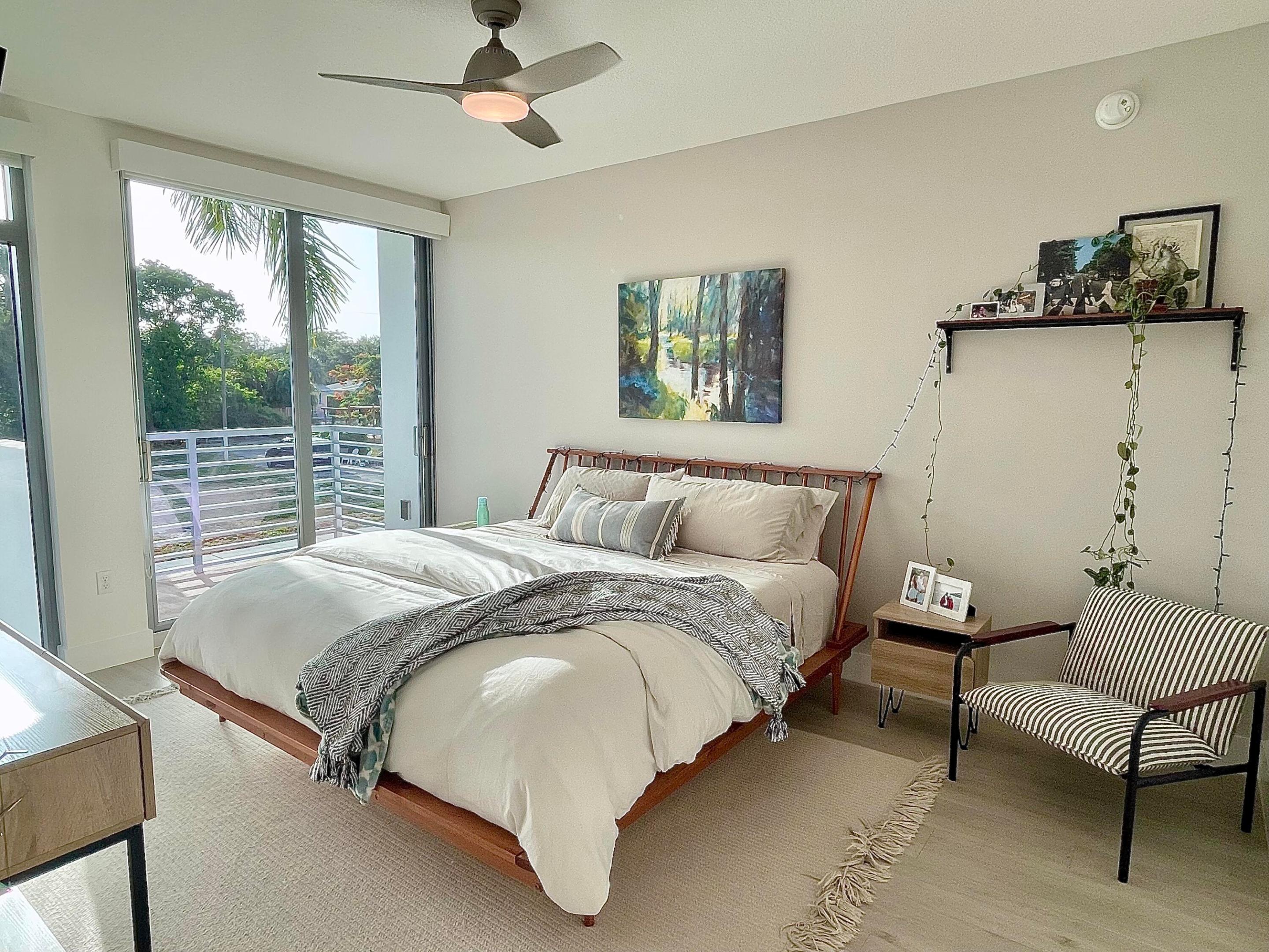 Bedroom with a wooden platform bed, striped lounge chair, and sliding doors to a tree-lined balcony.