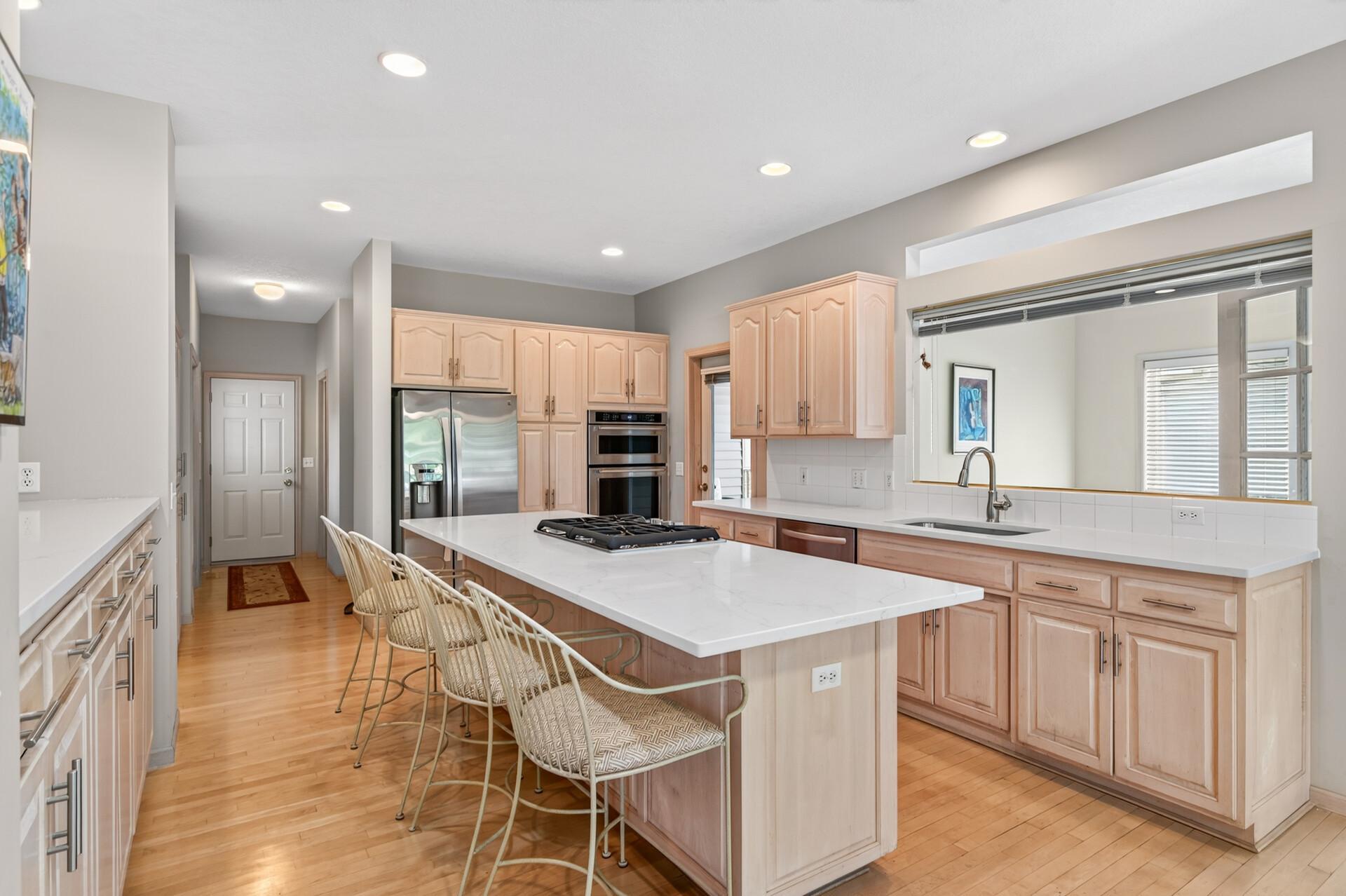 Kitchen with a large white island, gas cooktop, woven bar stools, and light wood cabinetry.