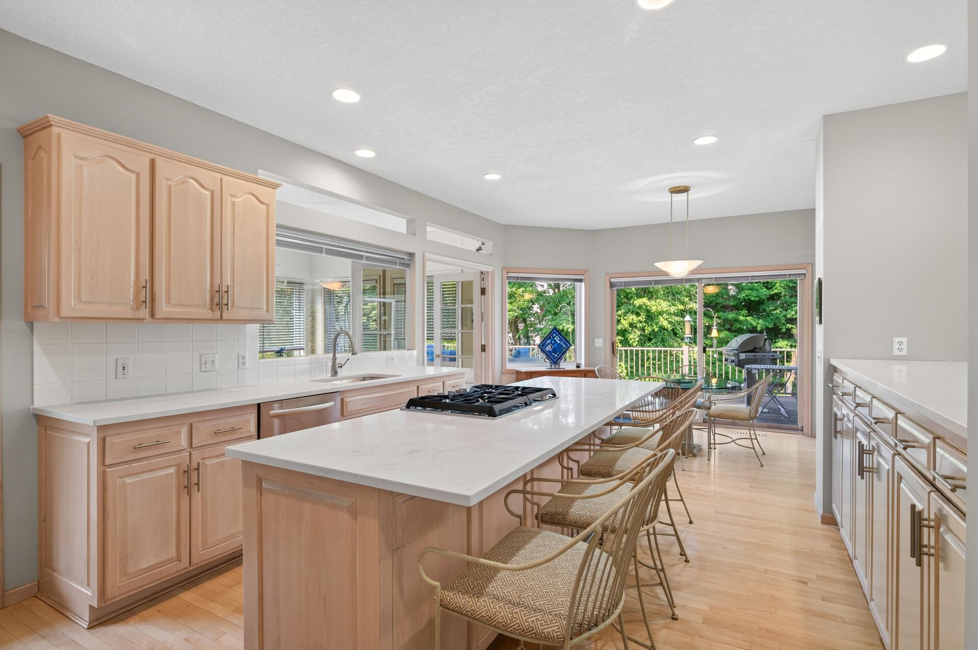 Kitchen with a large island, gas cooktop, light wood cabinets, and deck doors overlooking trees.