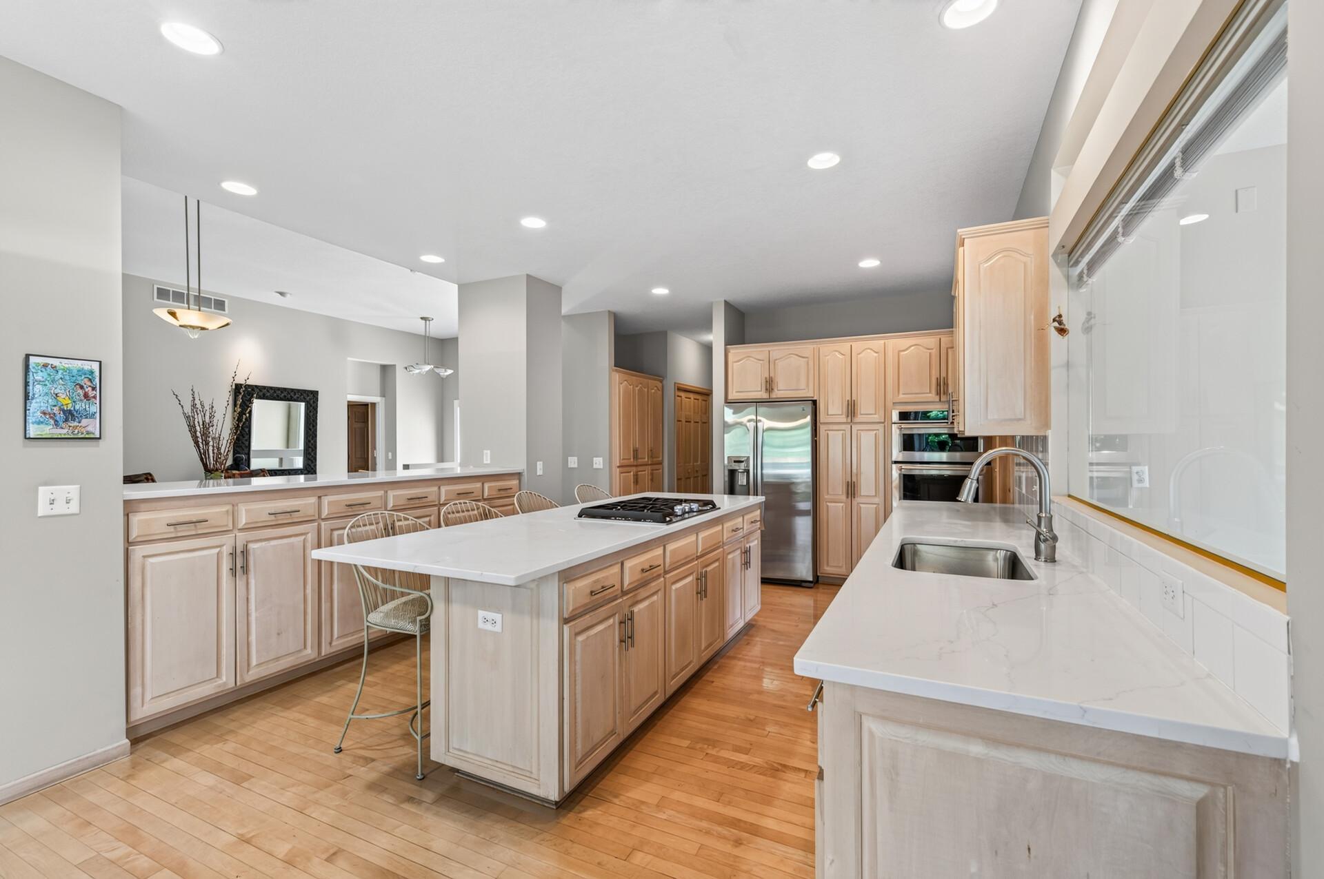 Spacious kitchen with a cooktop island, light wood cabinets, stainless steel appliances, and a sink beneath a wide window.
