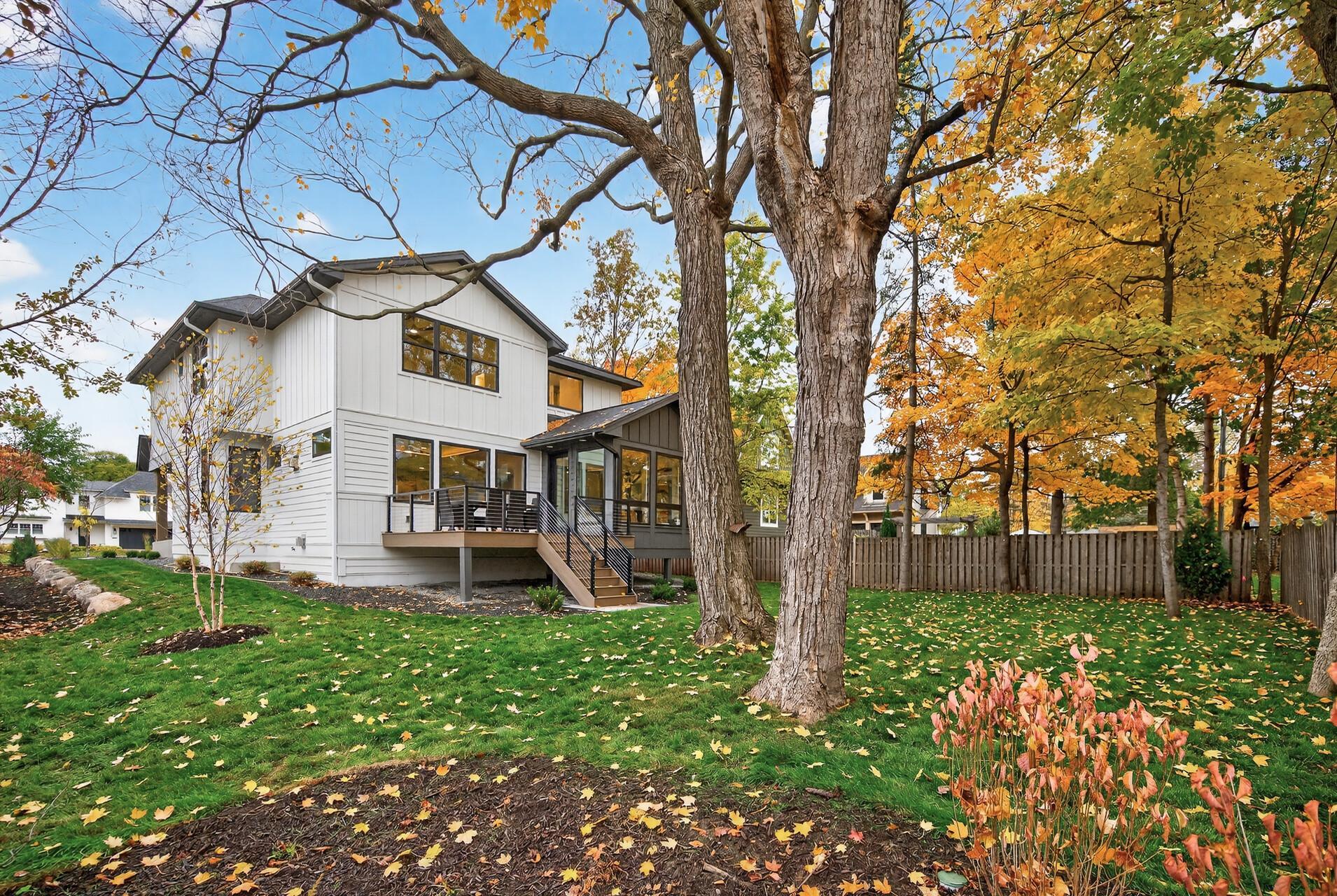 Two-story white-and-charcoal home beneath mature trees, with a raised deck and autumn foliage.