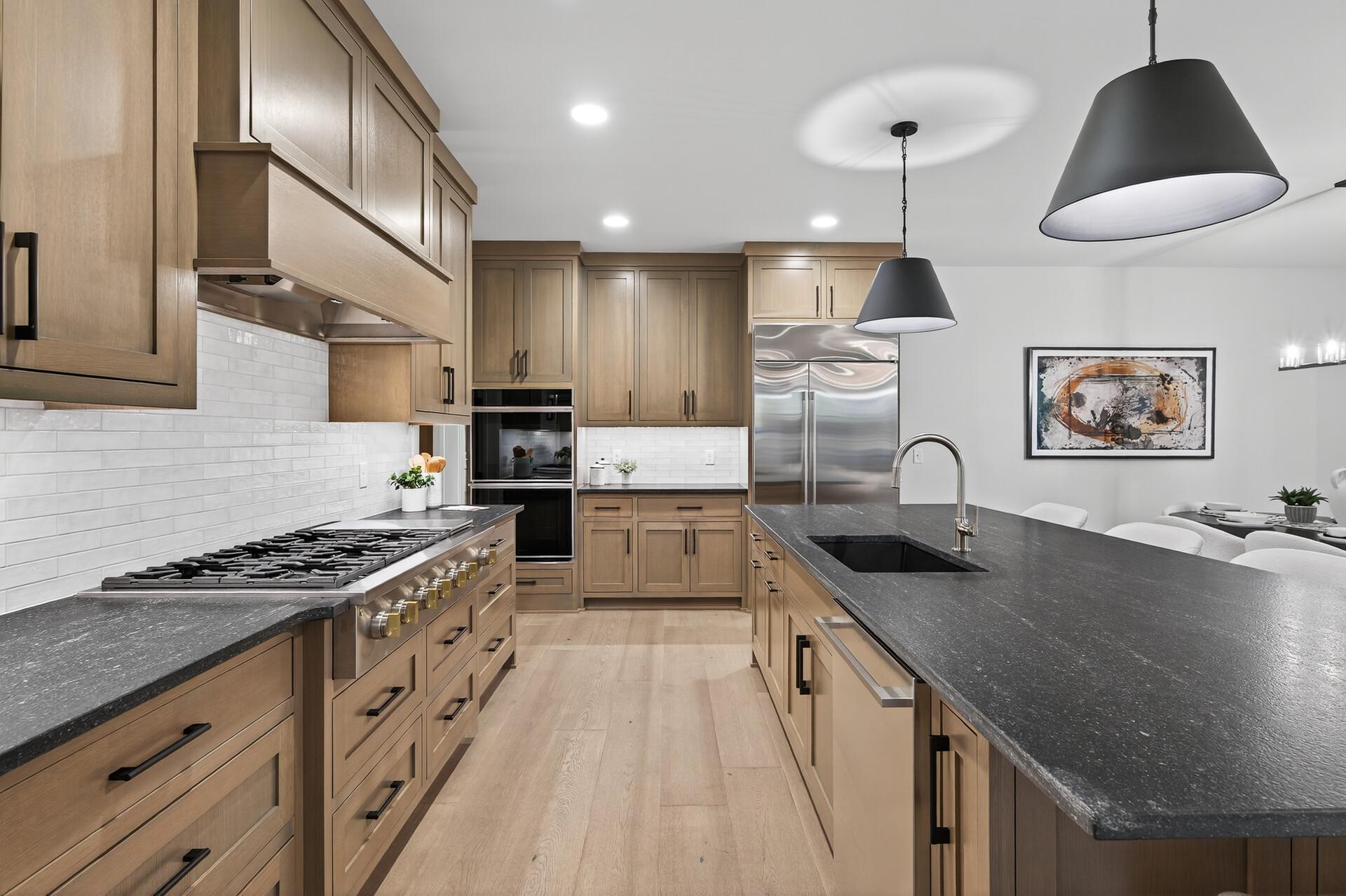 Modern kitchen with light wood cabinets, dark stone island, gas range, and stainless steel appliances.