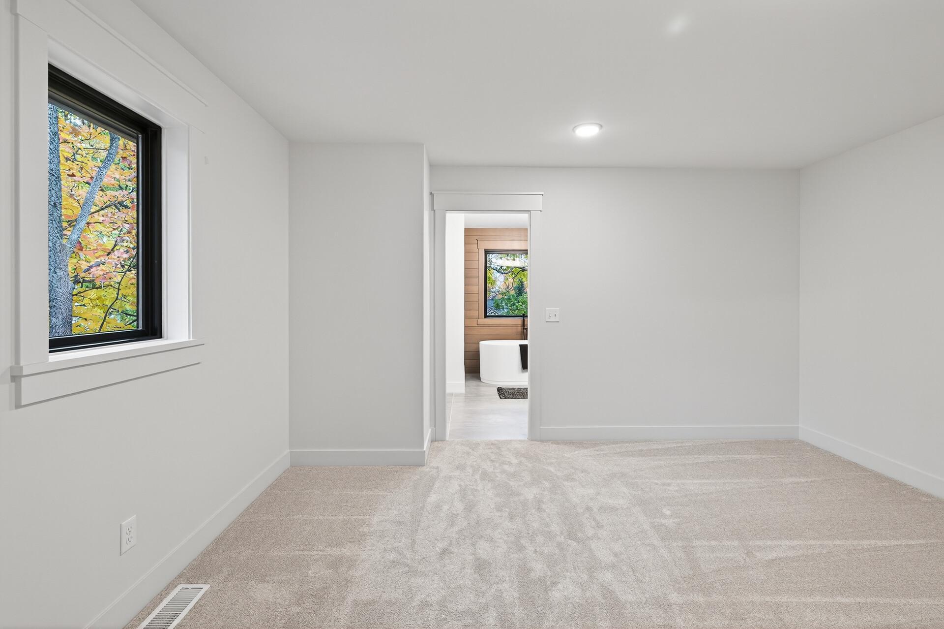 Empty carpeted bedroom with a large window and an open doorway to a bathroom with a freestanding tub.