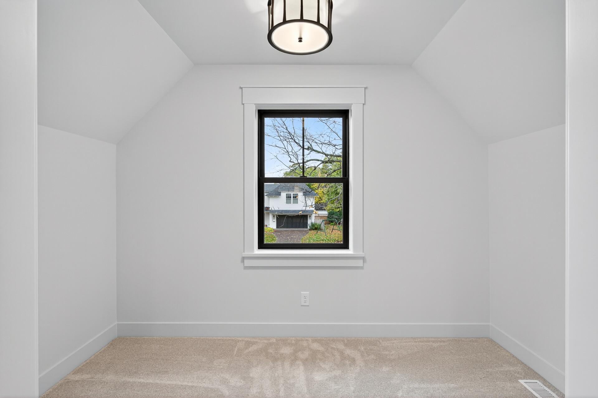 Empty carpeted bedroom with a sloped ceiling and window overlooking a neighboring home.