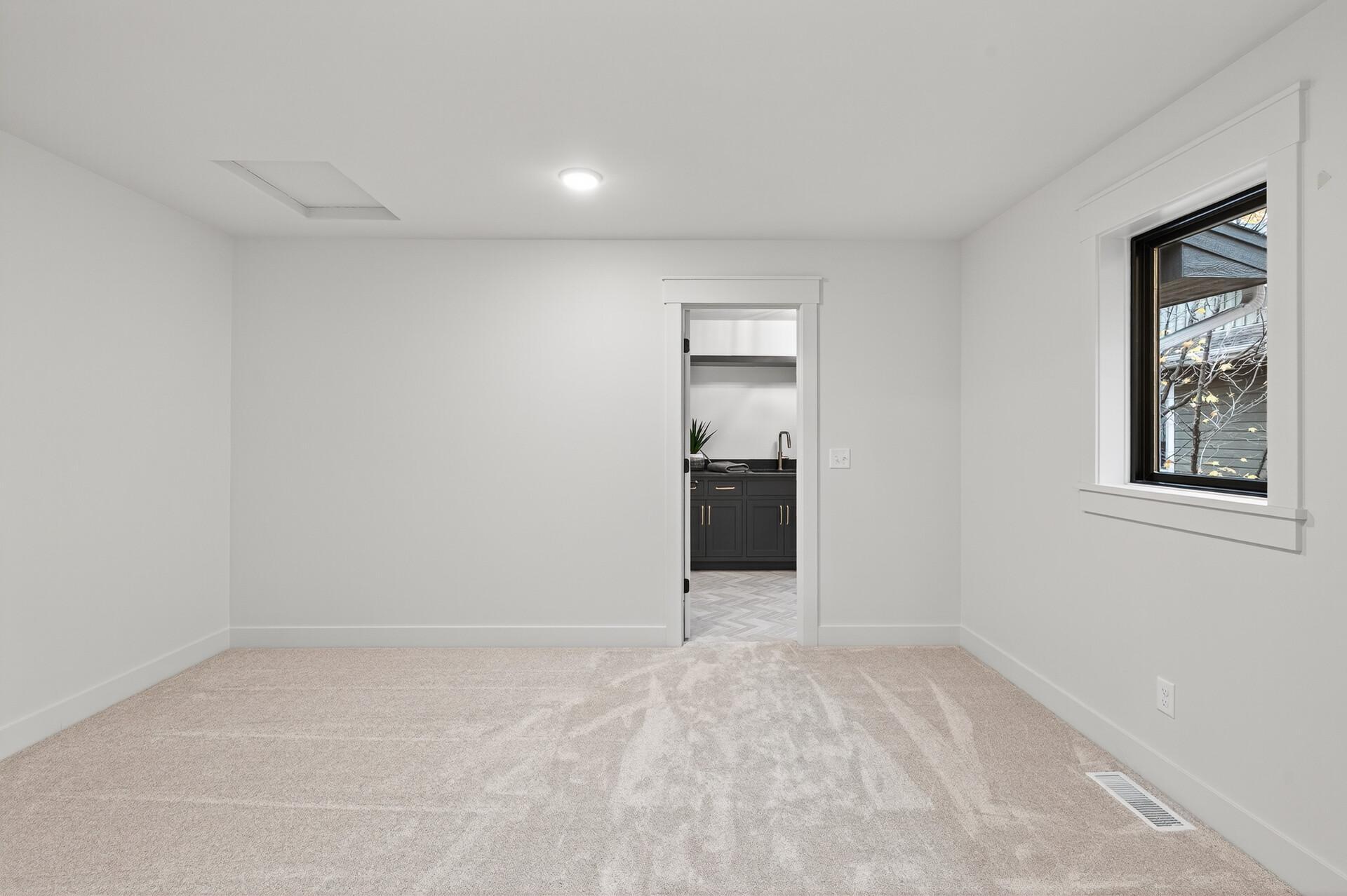 Vacant carpeted bedroom with a black-framed window and doorway to a room with dark cabinets and a sink.