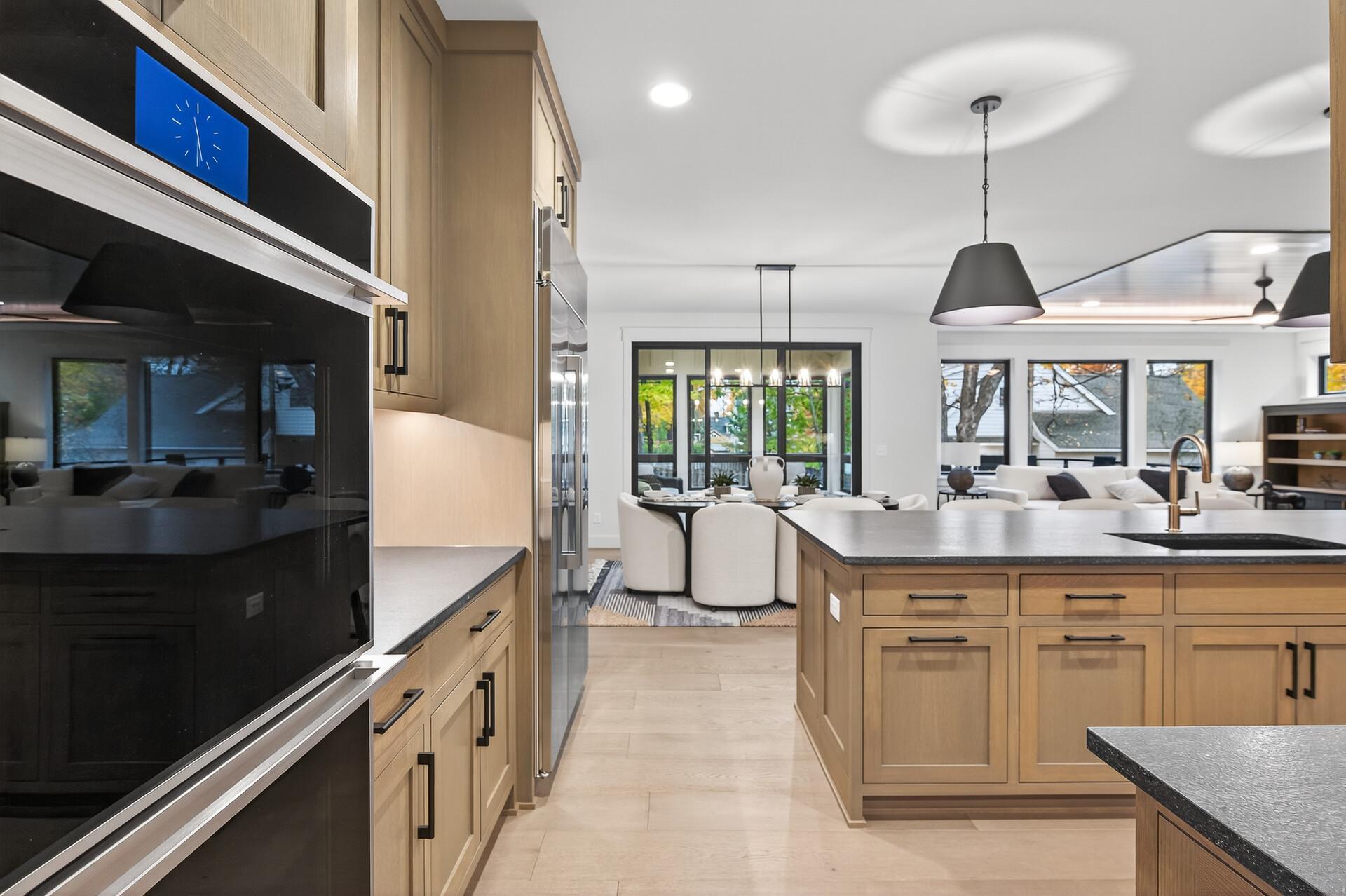 Open-plan kitchen with light wood cabinets, black ovens, a large island, and dining area by the windows.