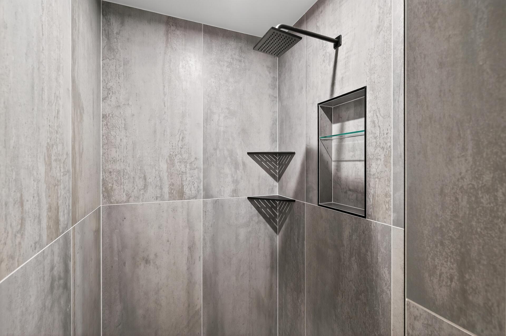 Walk-in shower with gray tile, black rainfall showerhead, corner shelves, and recessed niche.