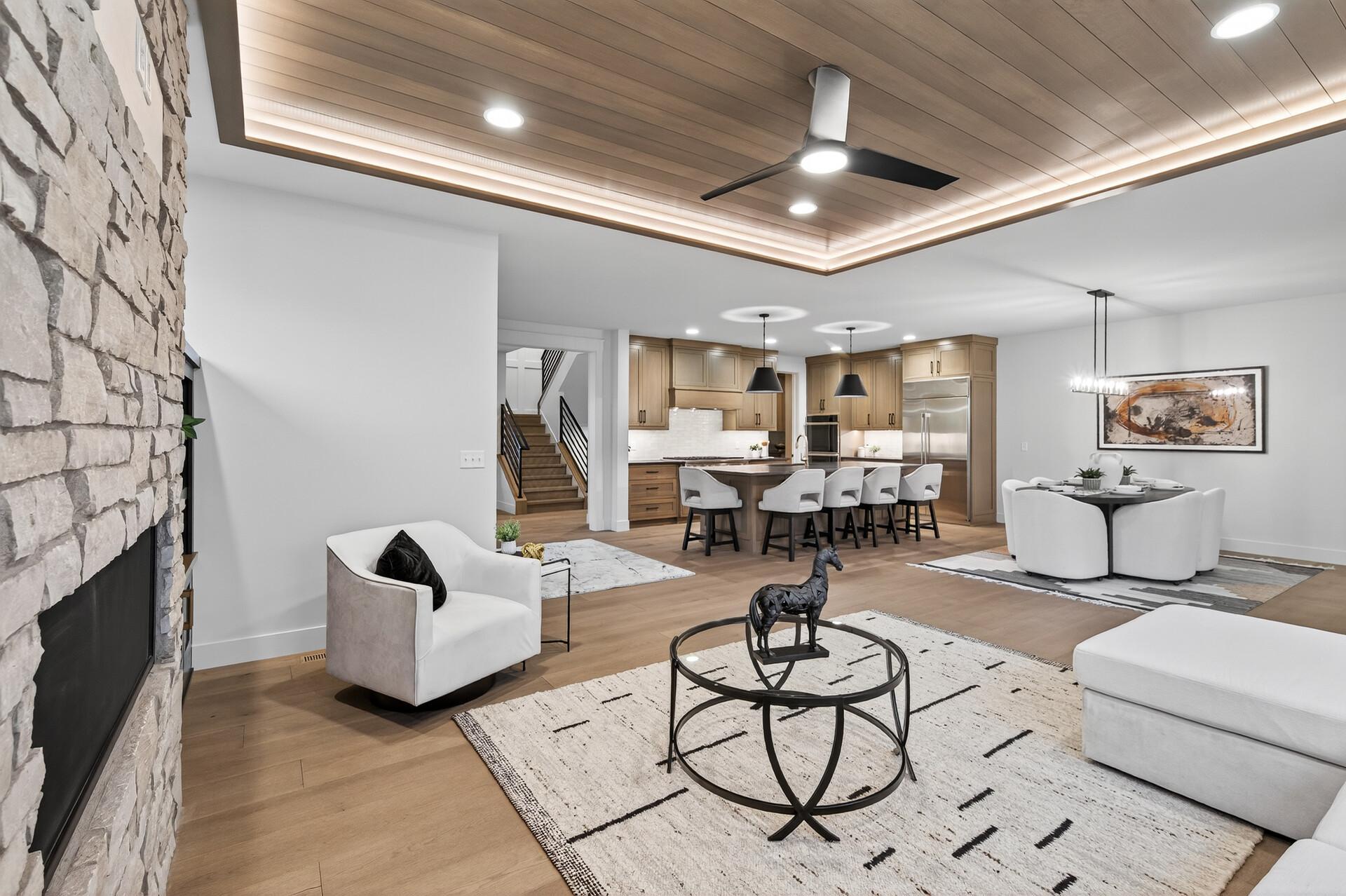Open living room with stone fireplace, wood-paneled ceiling, and adjoining kitchen and dining area.