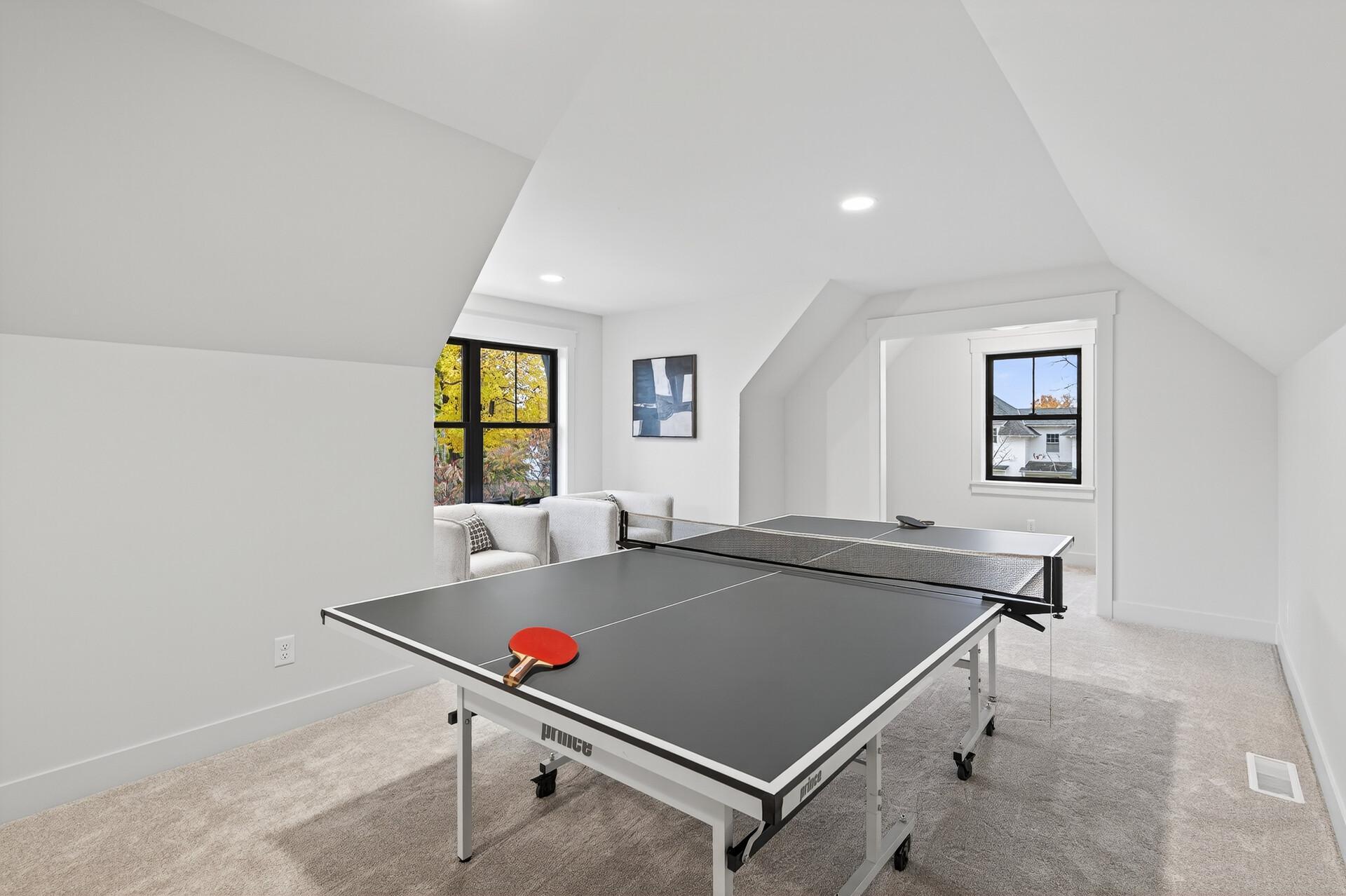 Attic recreation room with a ping-pong table, sloped ceilings, and windows overlooking autumn trees.