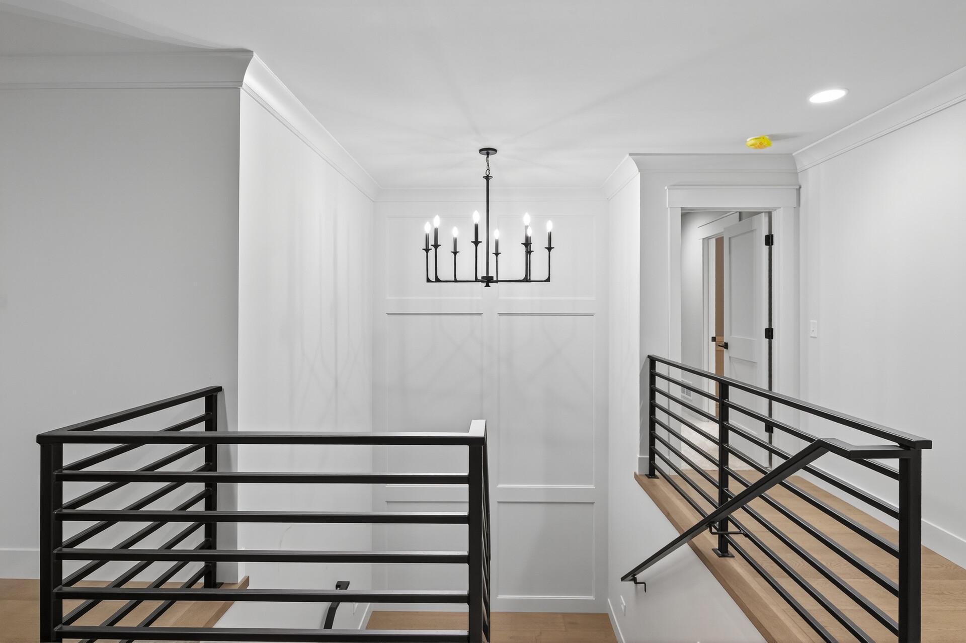 Bright stair landing with black metal railings, light wood treads, and a black chandelier.