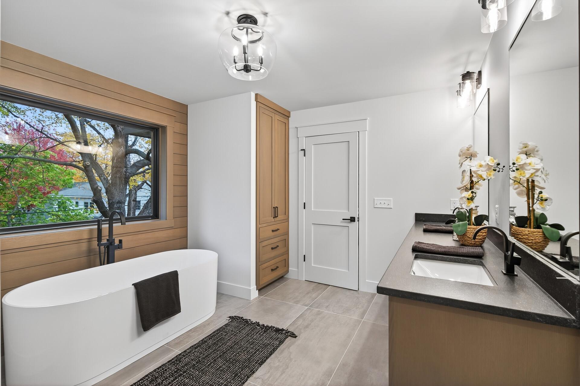 Modern bathroom with a freestanding tub beneath a large window and a dark stone vanity.