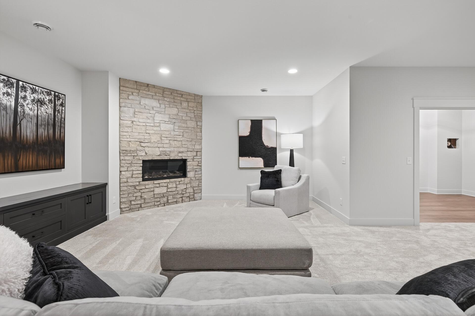 Living room with a stone fireplace, pale carpet, upholstered ottoman, and dark built-in media cabinet.