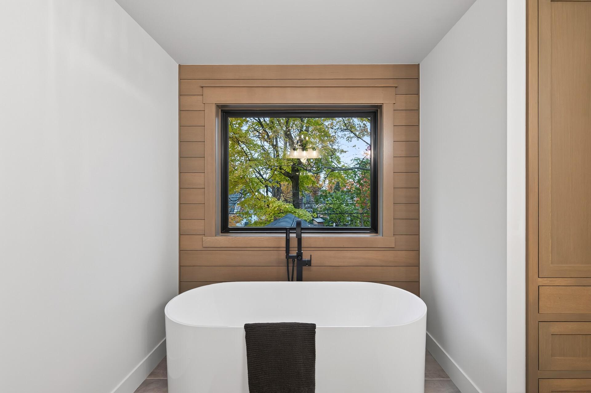 Bathroom with a freestanding soaking tub beneath a wood-framed window overlooking leafy trees.