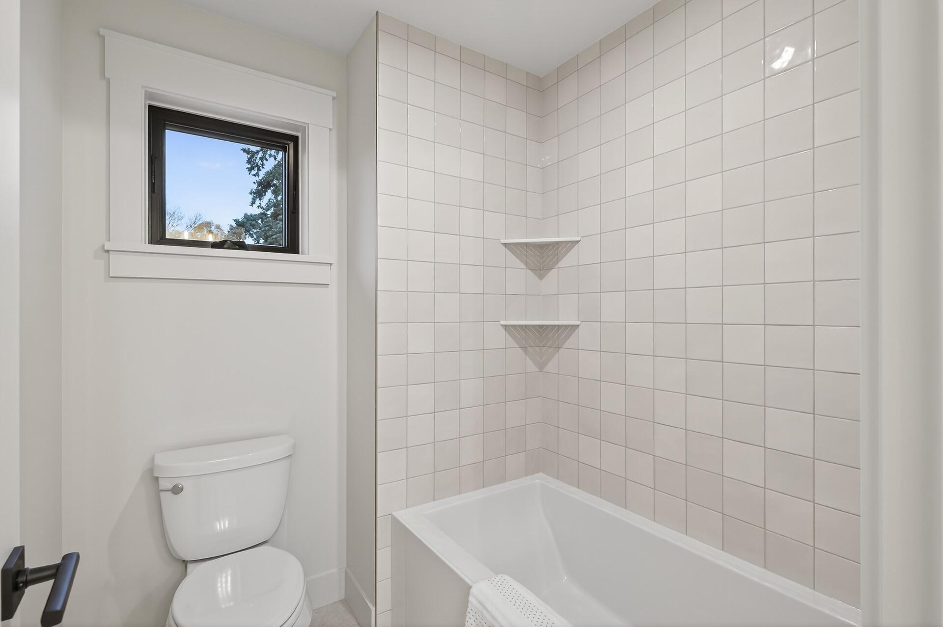 Bathroom with a white bathtub, tiled shower wall, corner shelves, toilet, and window.