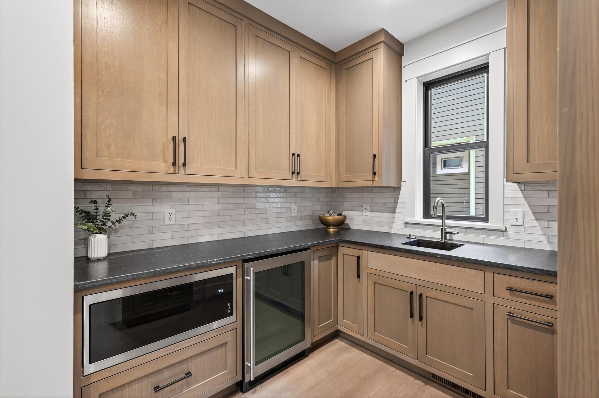 Kitchen wet bar with light wood cabinets, dark counters, subway tile backsplash, sink, microwave, and beverage refrigerator.