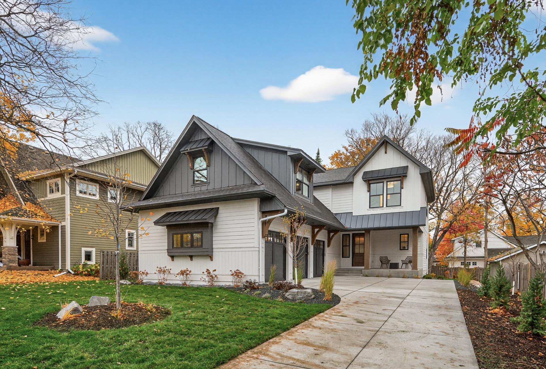 Modern farmhouse with steep gables, a covered porch, and a concrete driveway beneath autumn trees.