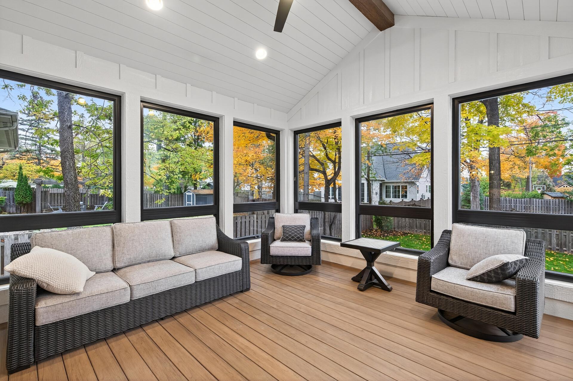 Furnished sunroom with wicker seating, vaulted ceiling, and large windows overlooking autumn foliage.