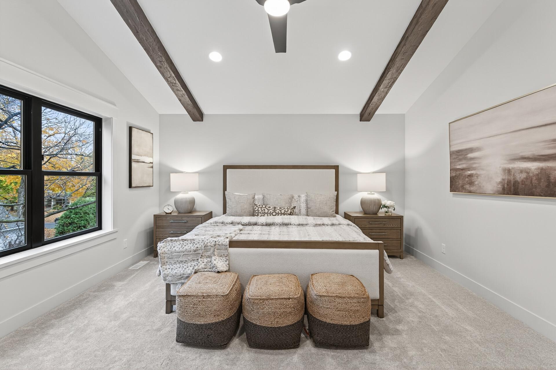 Staged bedroom with exposed wood ceiling beams, a centered bed, and large windows overlooking autumn trees.