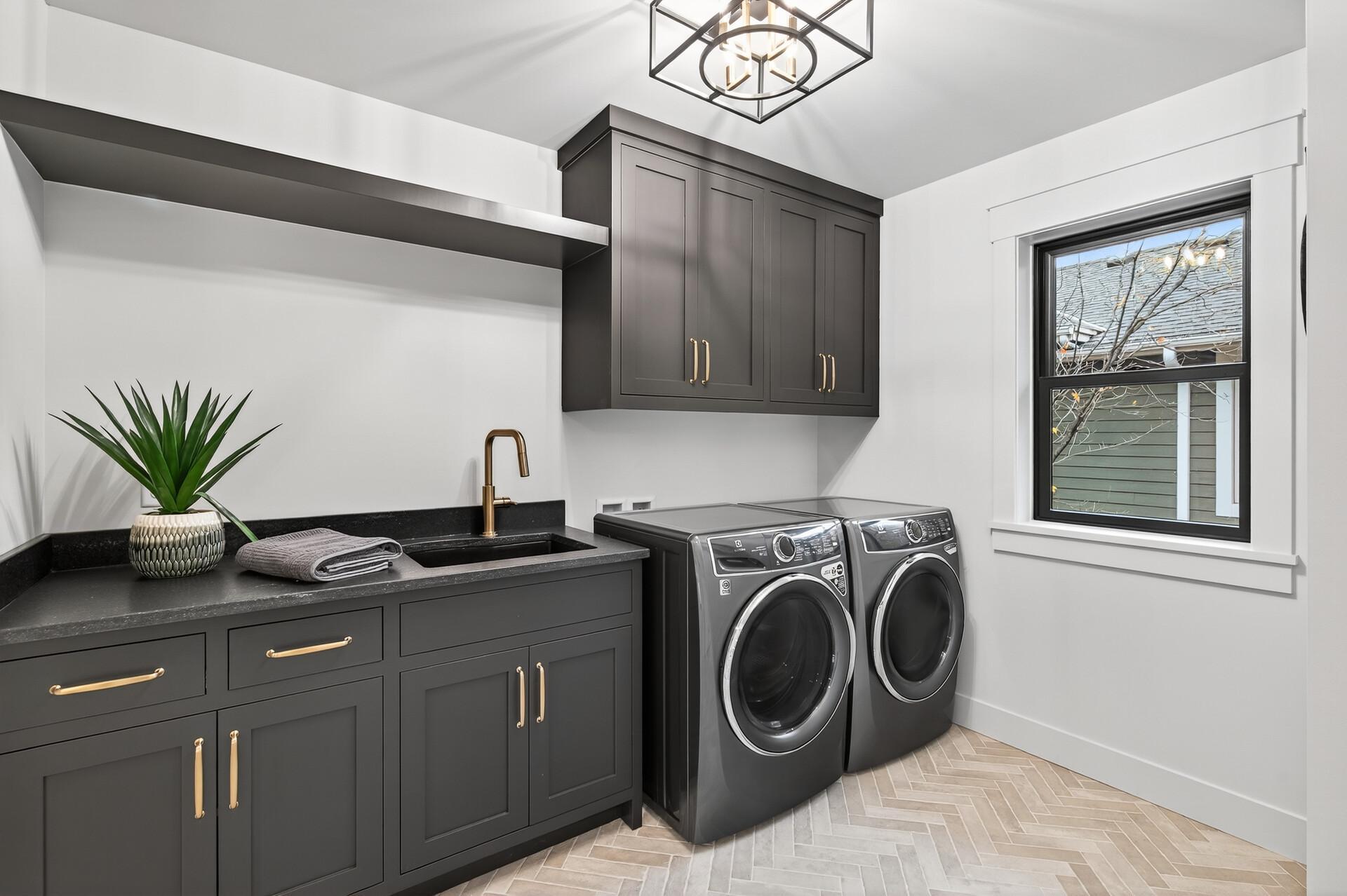 Laundry room with charcoal cabinets, front-loading washer and dryer, utility sink, and herringbone tile floor.