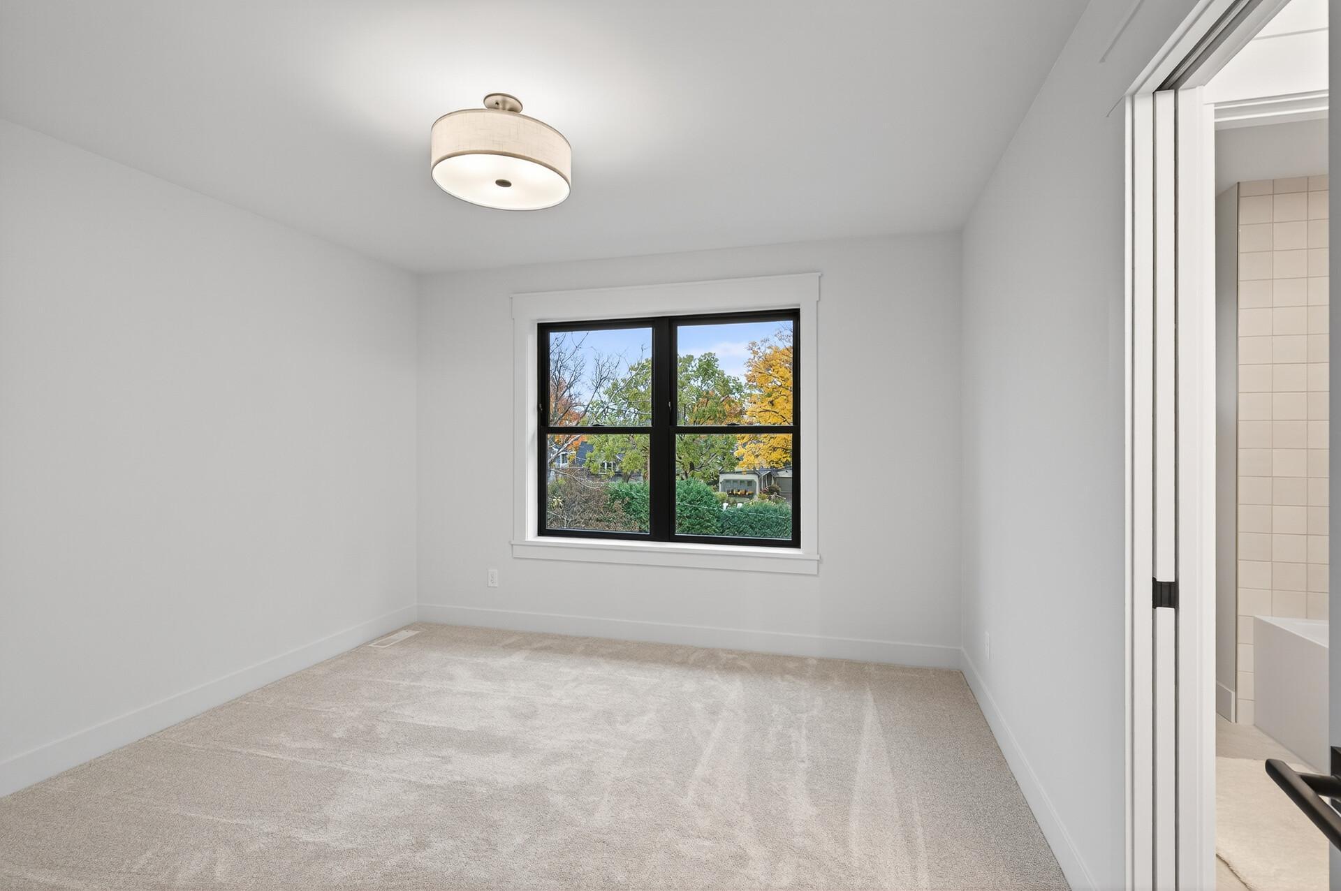 Empty bedroom with beige carpet and a black-framed window overlooking autumn trees.