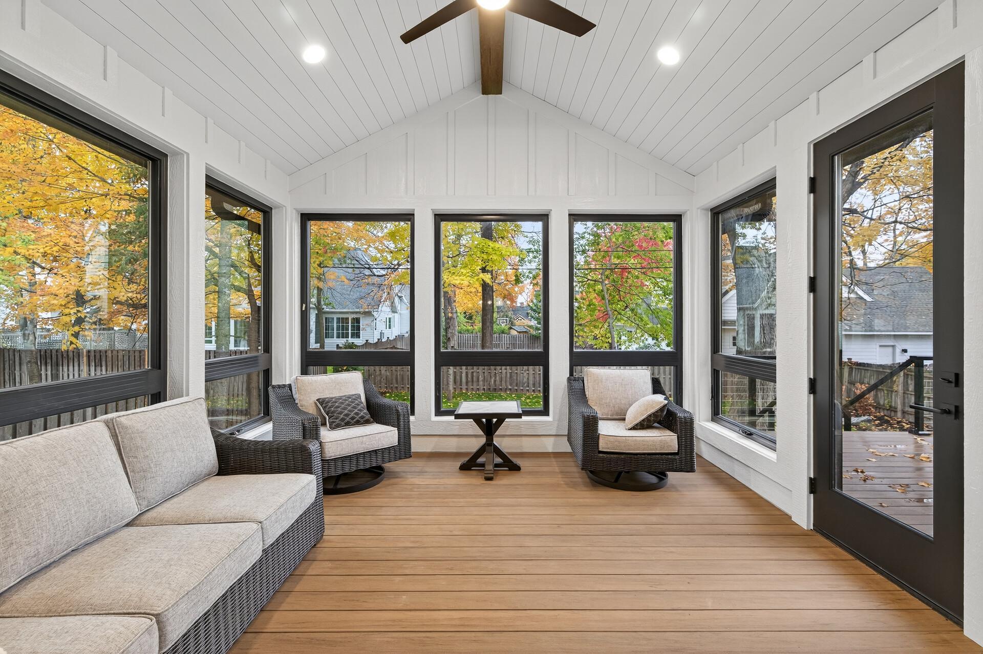 Enclosed sunroom with wicker seating, vaulted white ceiling, and large windows overlooking autumn trees.