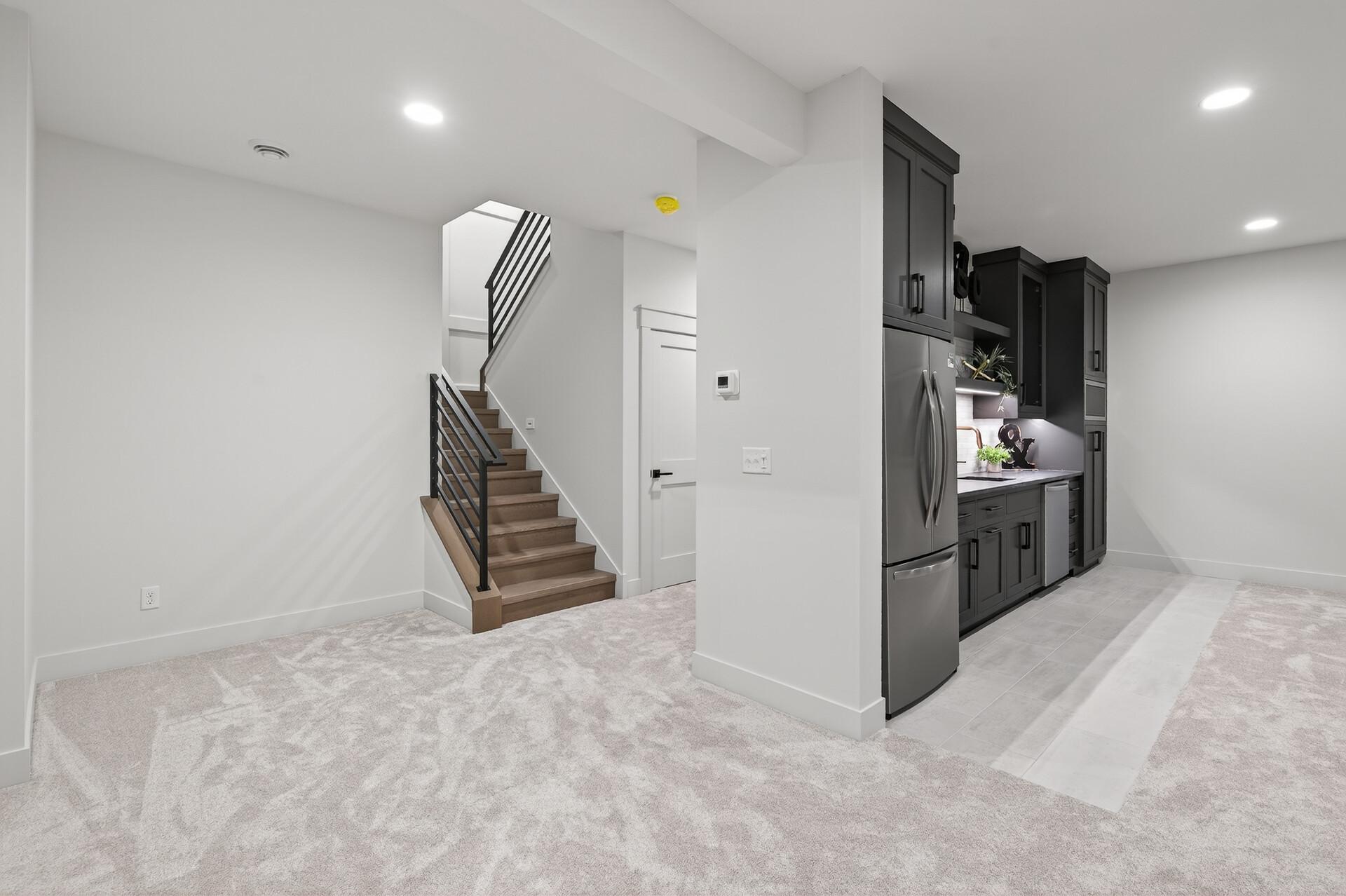 Open kitchen and carpeted area beside a wood staircase with black railings.