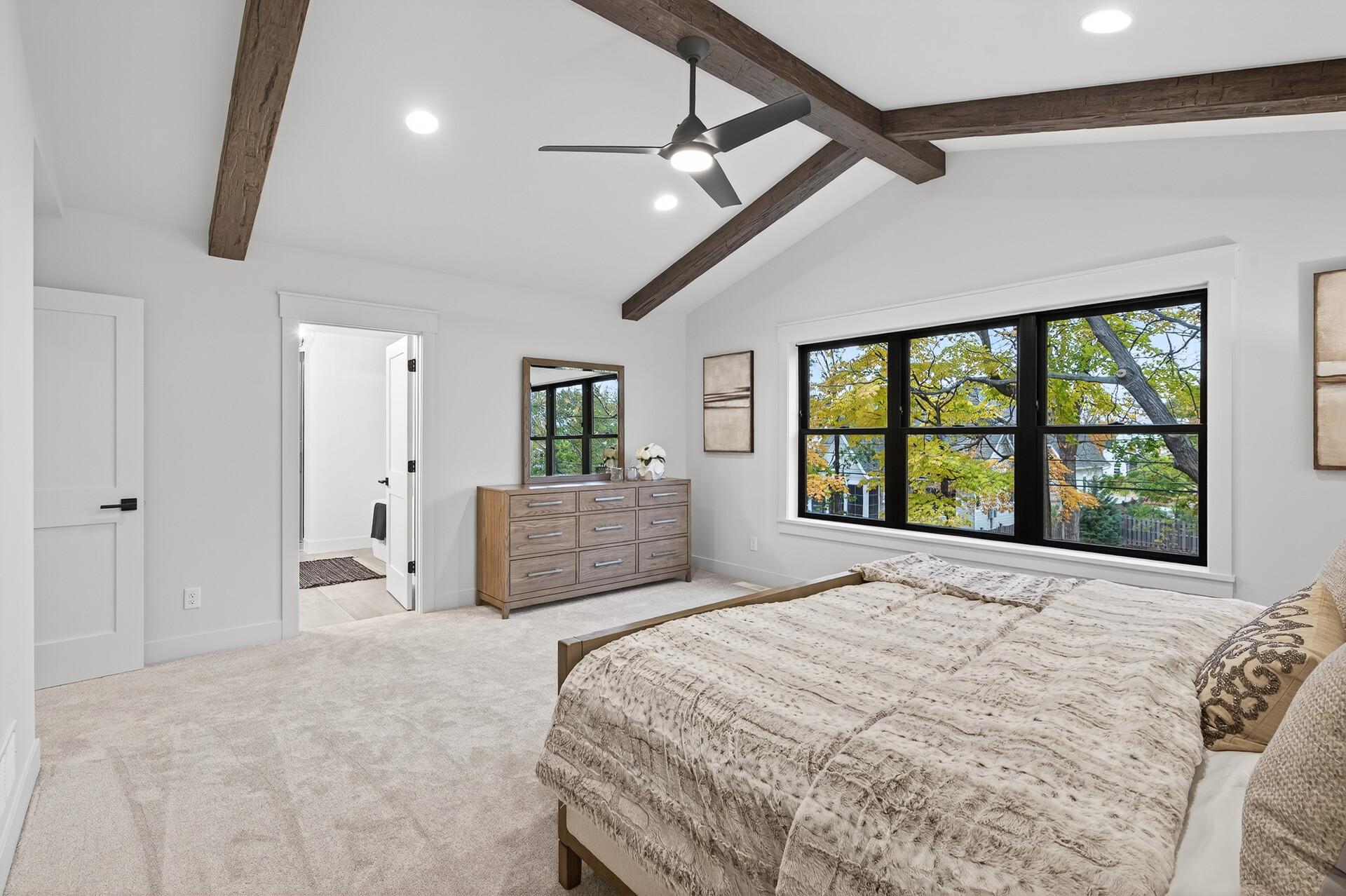 Bedroom with exposed ceiling beams, a patterned bed, and large windows overlooking trees.