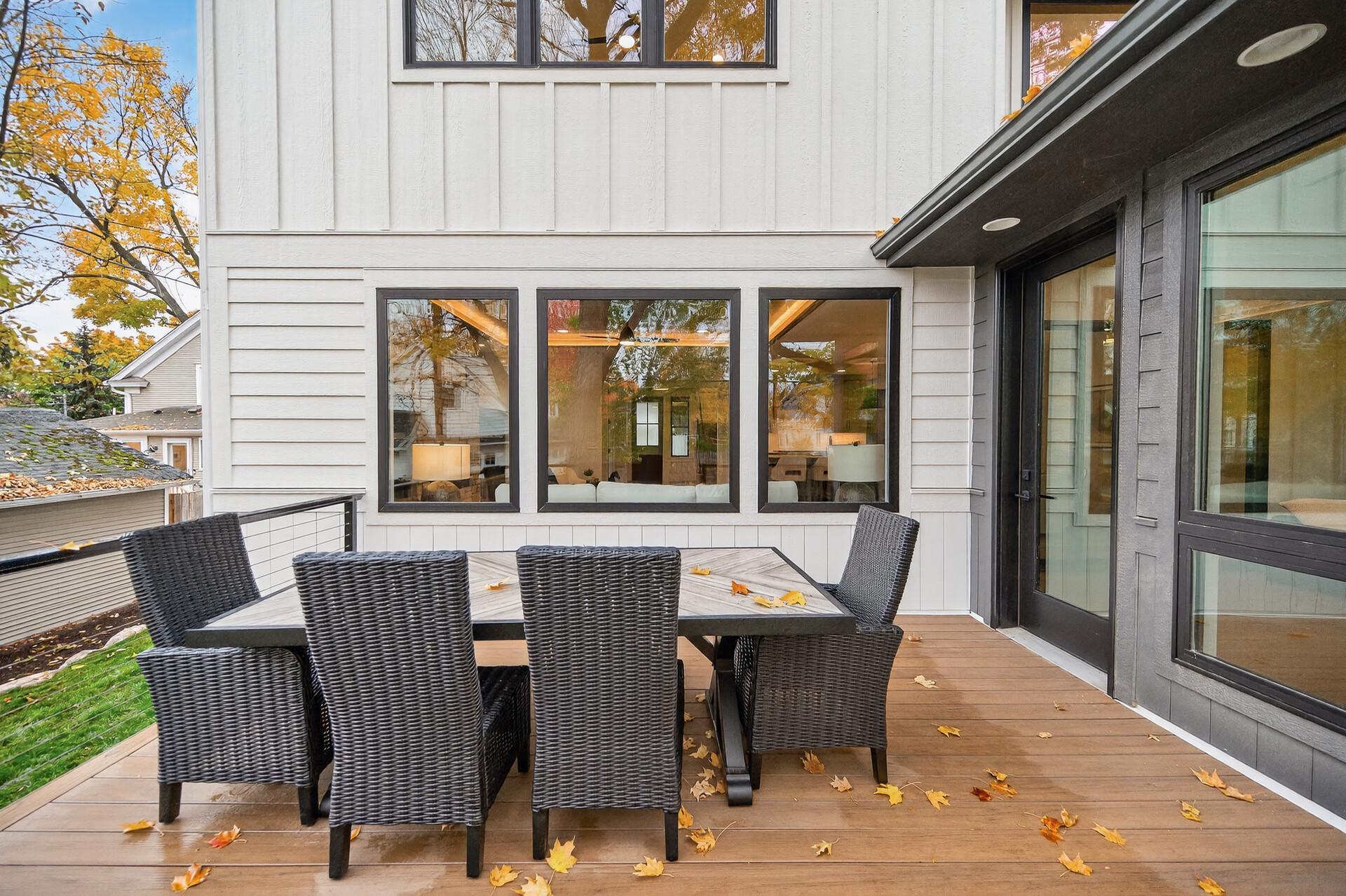 Outdoor deck with a woven dining set, large windows, and autumn leaves on the wood flooring.