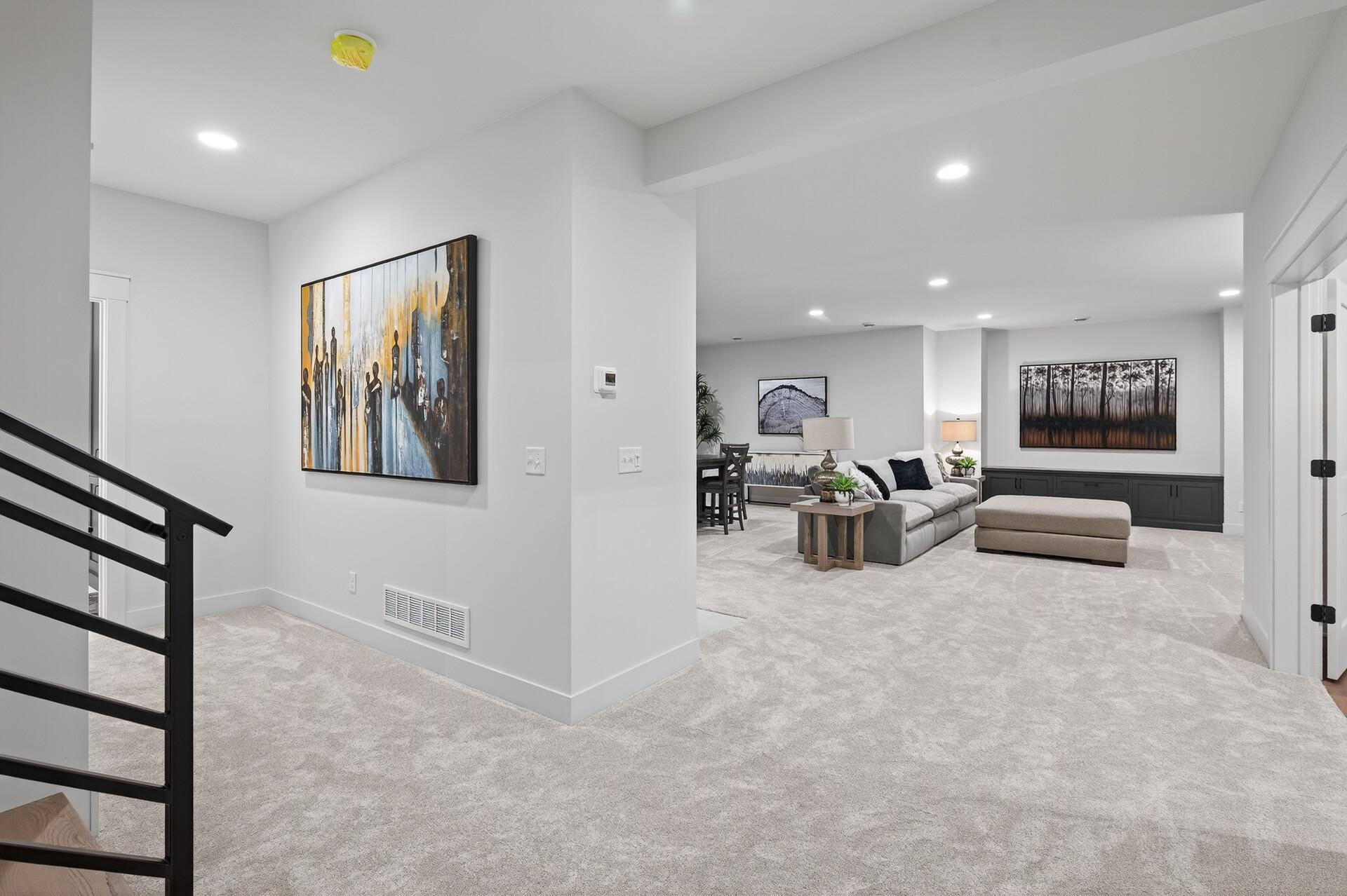 Open living area with gray sectional and ottoman, dining space beyond, and black stair railing.