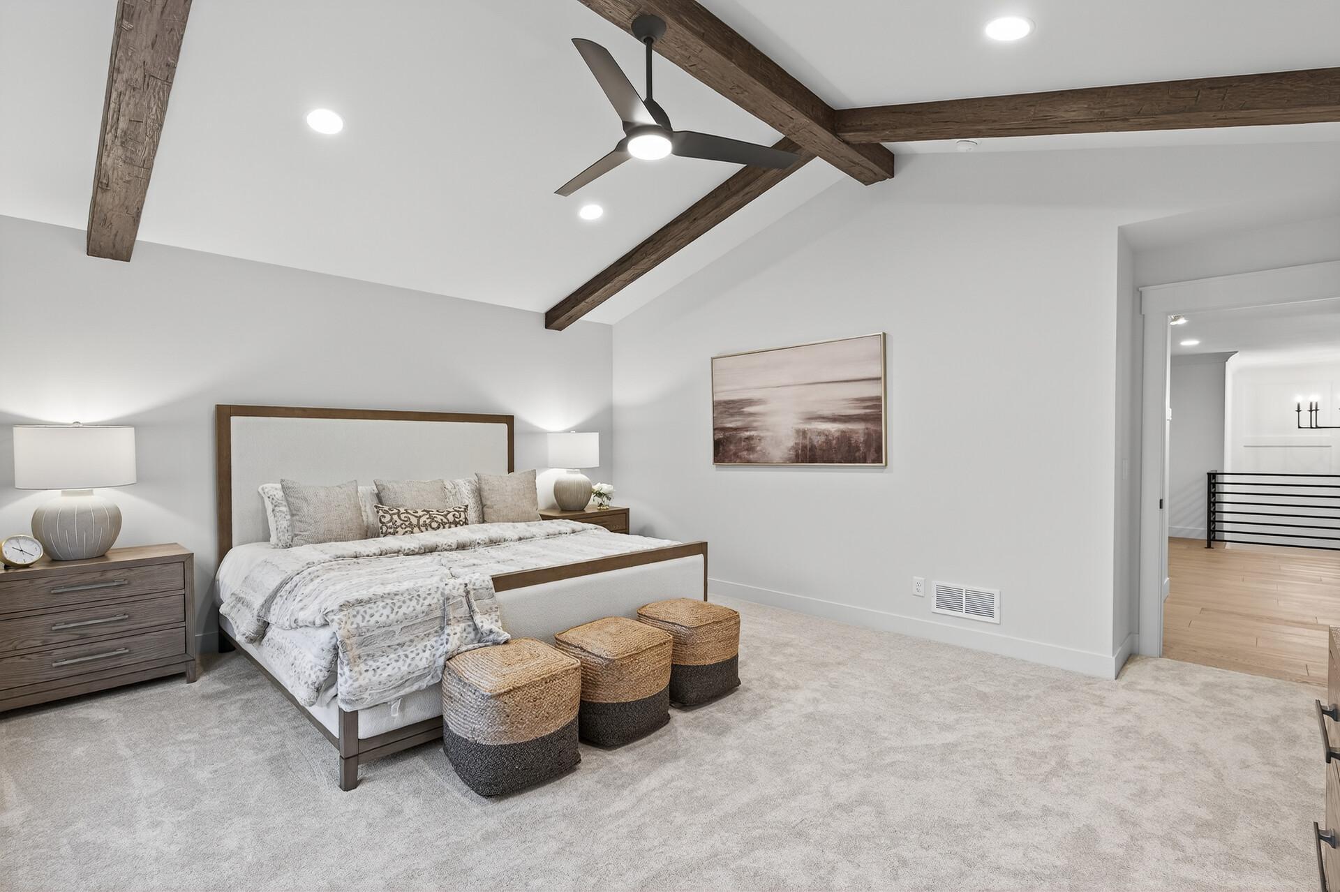 Bedroom with exposed dark ceiling beams, pale bedding, and woven ottomans at the foot of the bed.