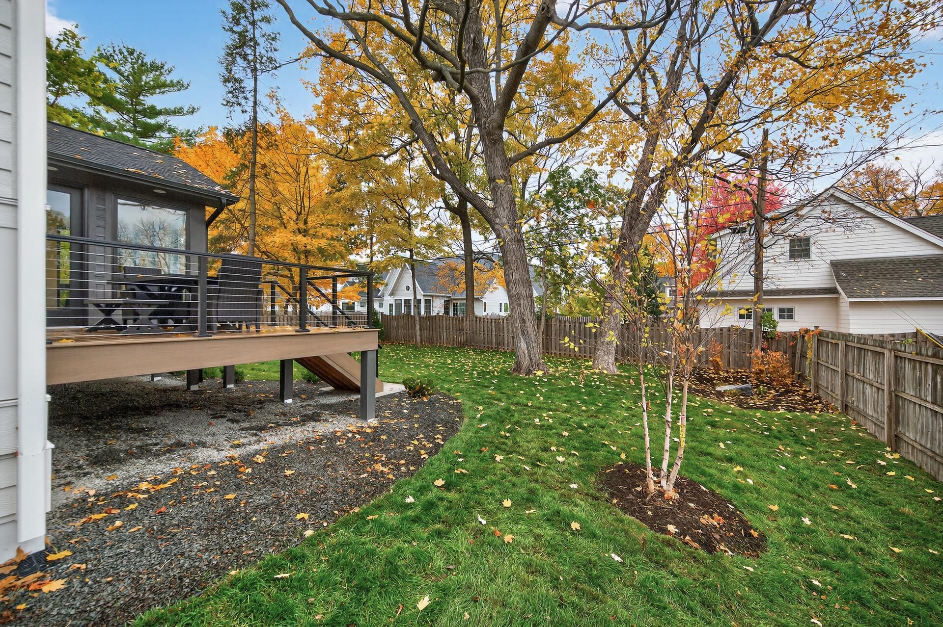 Fenced backyard with a raised deck beneath large autumn trees.