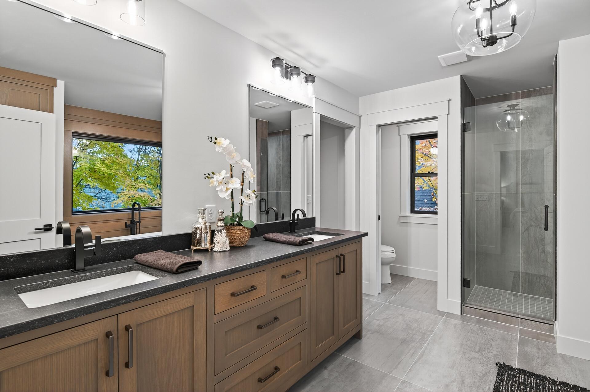 Bathroom with a double-sink wood vanity, dark countertop, and glass shower; fall foliage and water show through the windows.