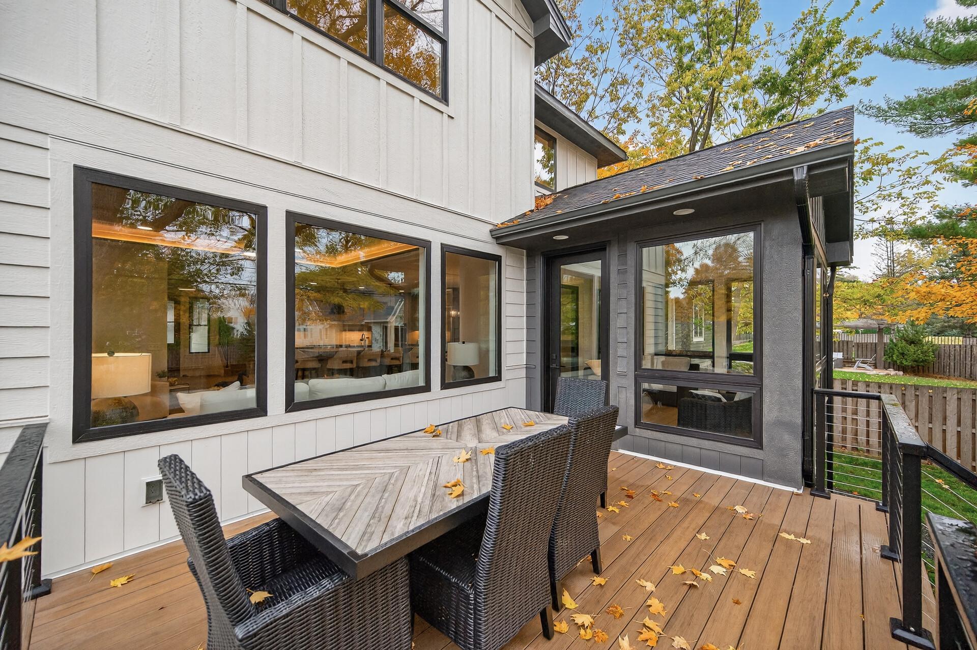 Wooden deck with wicker dining set beside a white house with dark trim and large windows.