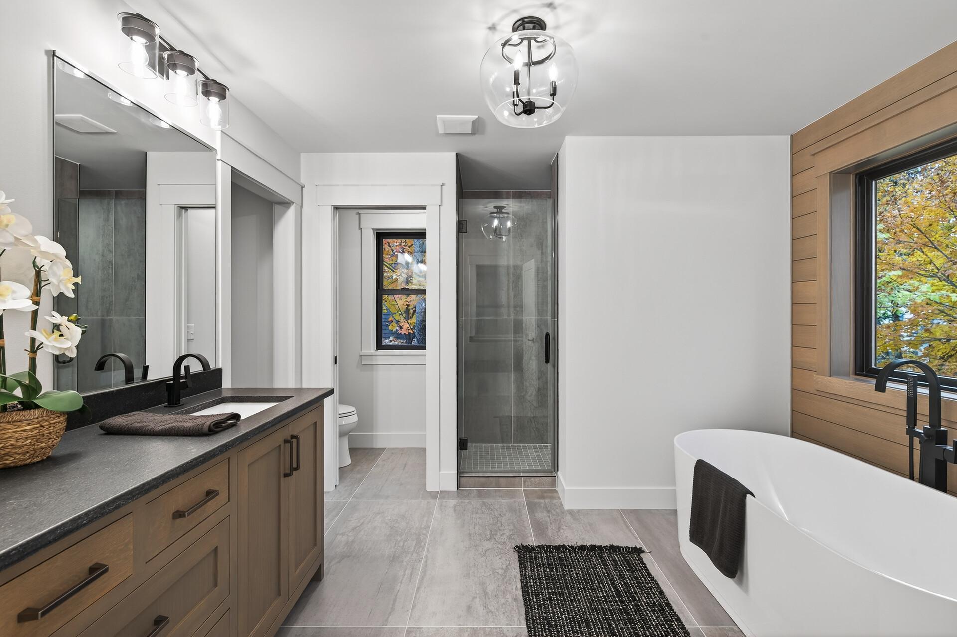 Bathroom with a wood vanity, glass shower, and freestanding tub beside a window with autumn foliage.
