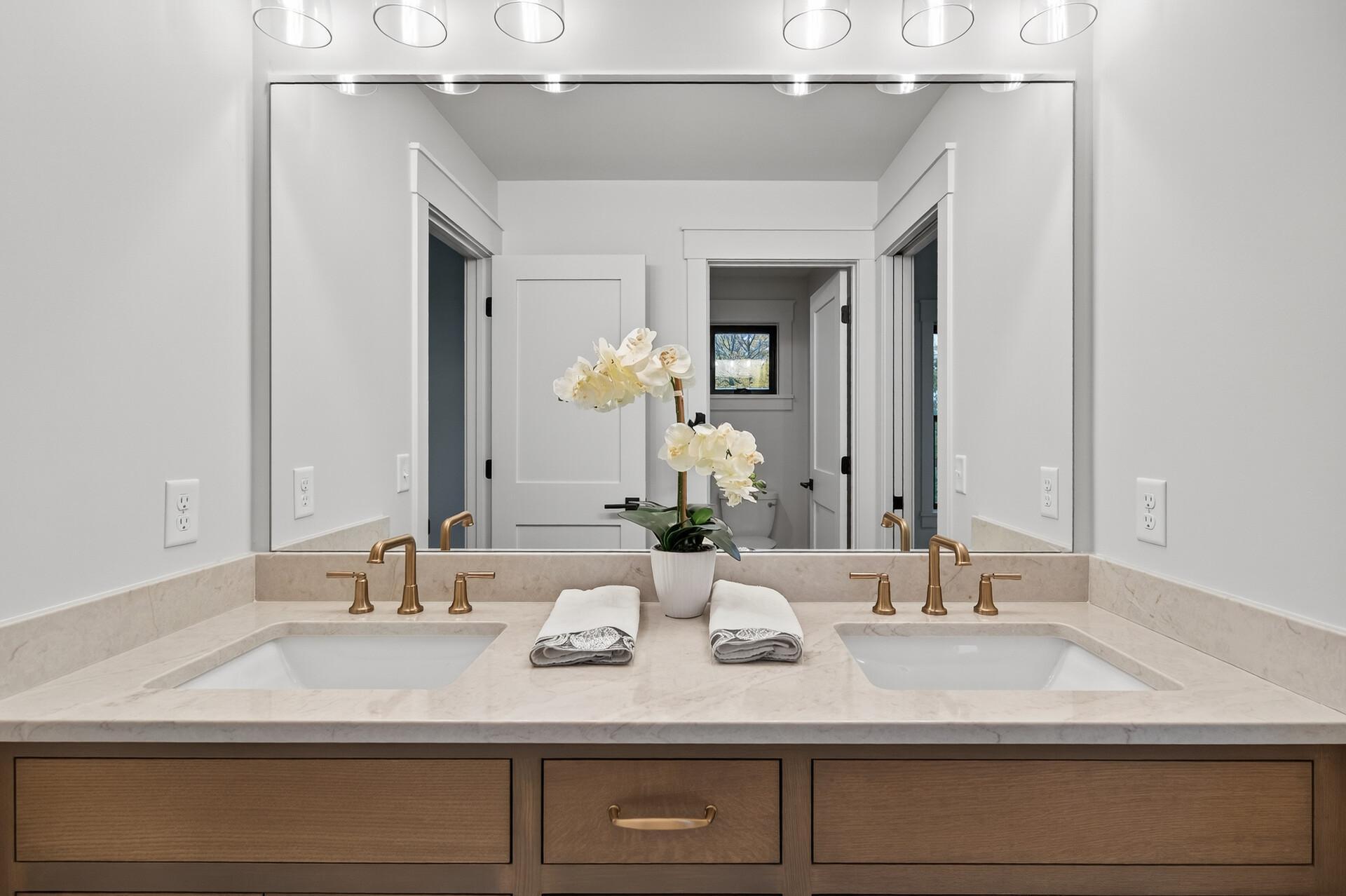 Bathroom double vanity with brass faucets, pale stone counters, and an orchid between the sinks.