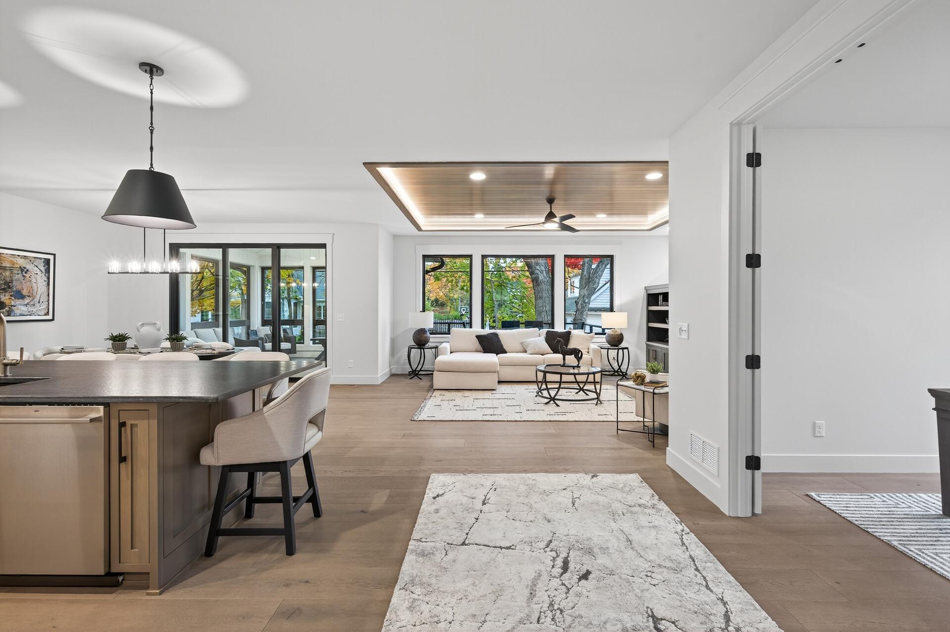 Open-plan kitchen and living room with a dark island, white seating, and windows overlooking autumn trees.