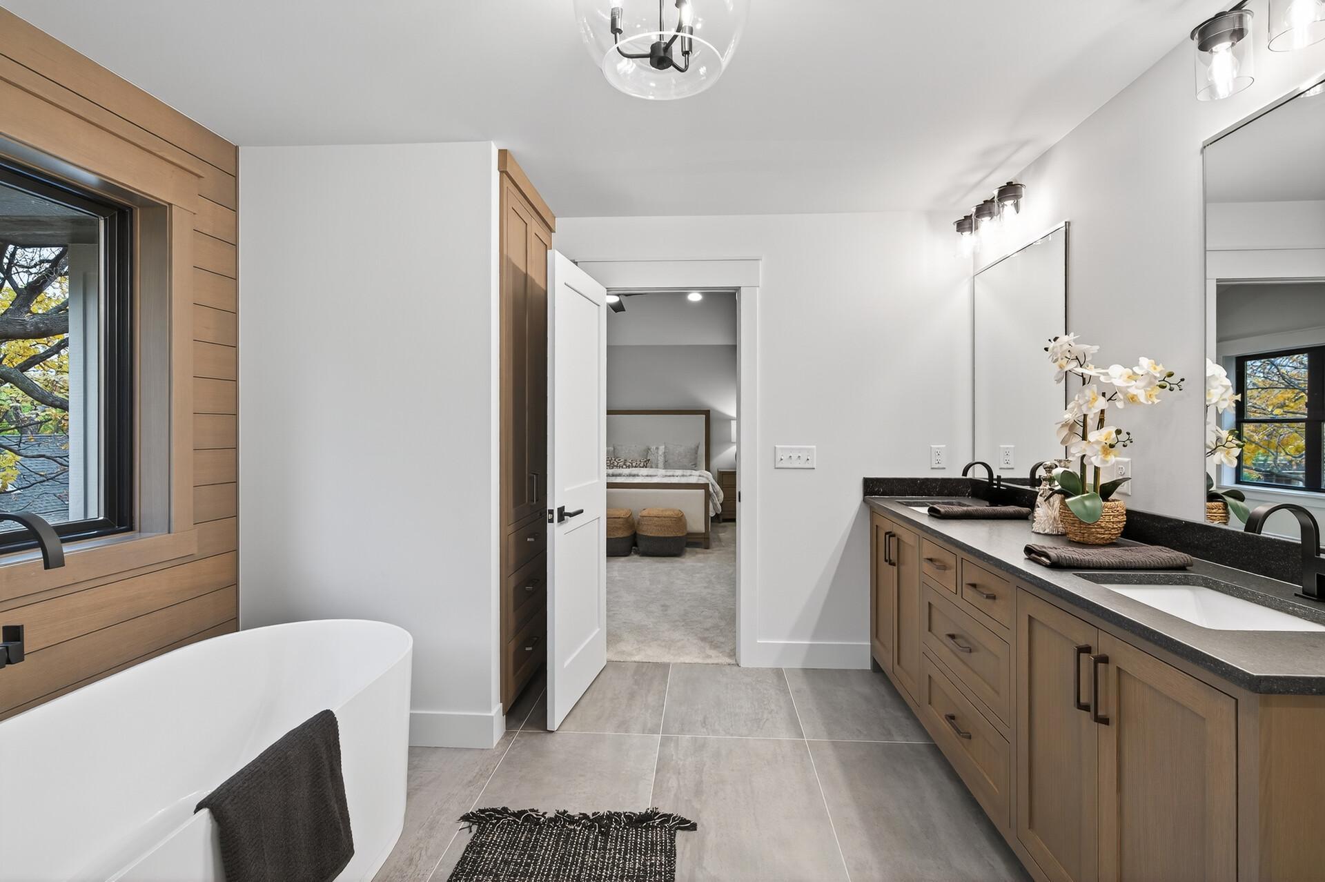 Modern bathroom with a freestanding tub, wood vanity, dark countertop, and window overlooking autumn trees.