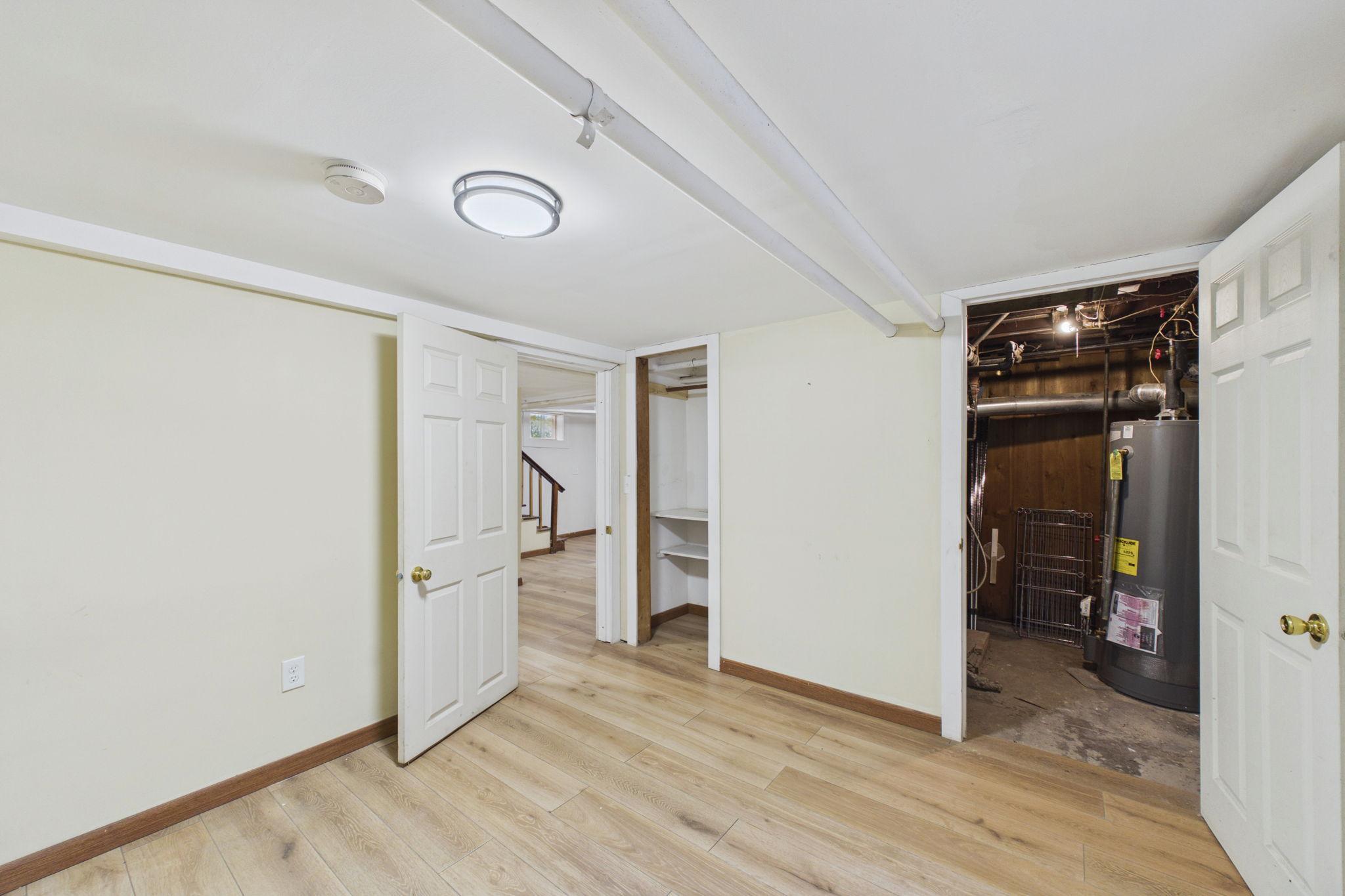 Basement room with light wood-look flooring, open storage closet, and utility area with a water heater.