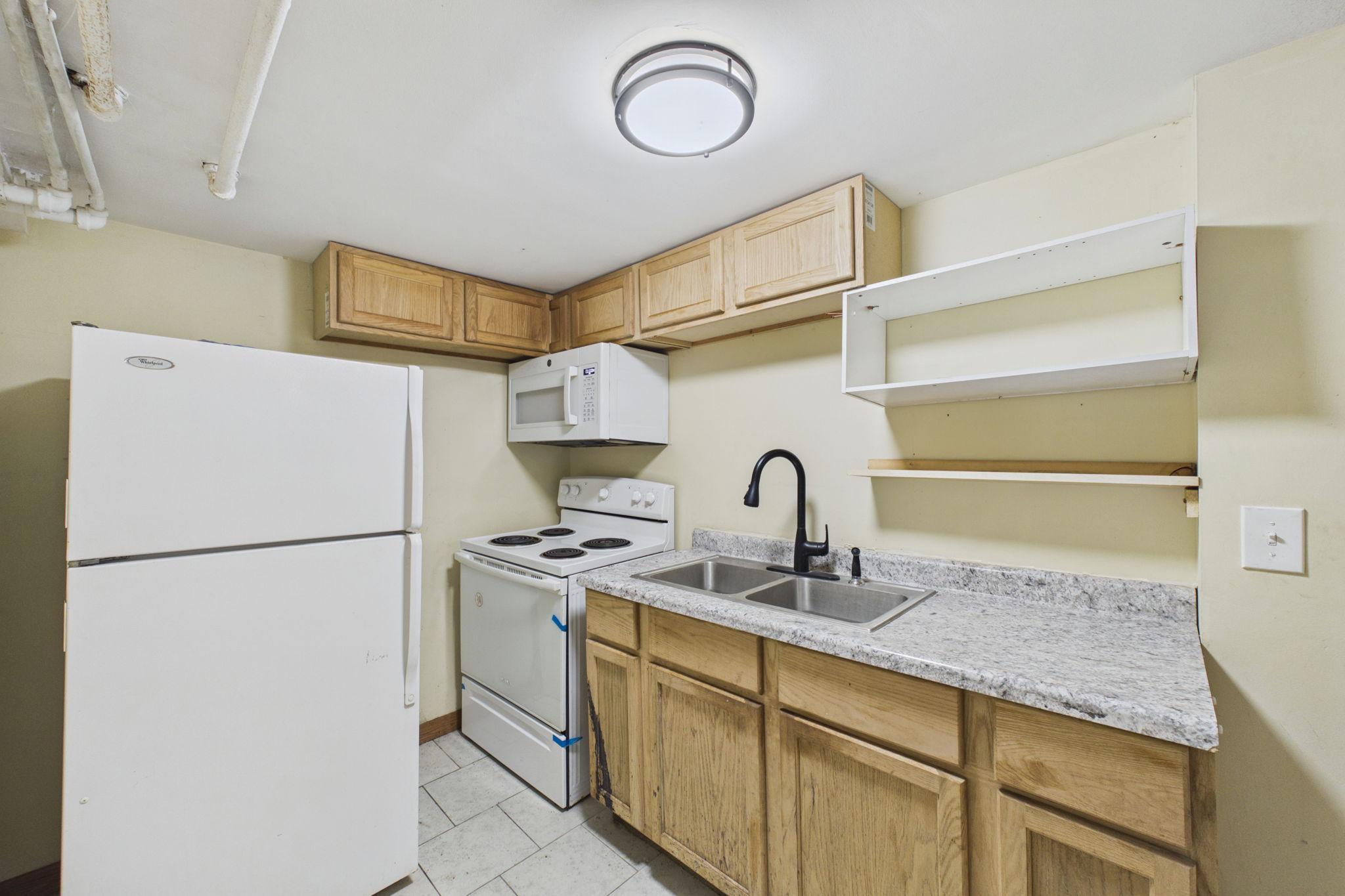 Compact kitchen with wood cabinets, speckled counters, a double sink, and white appliances.