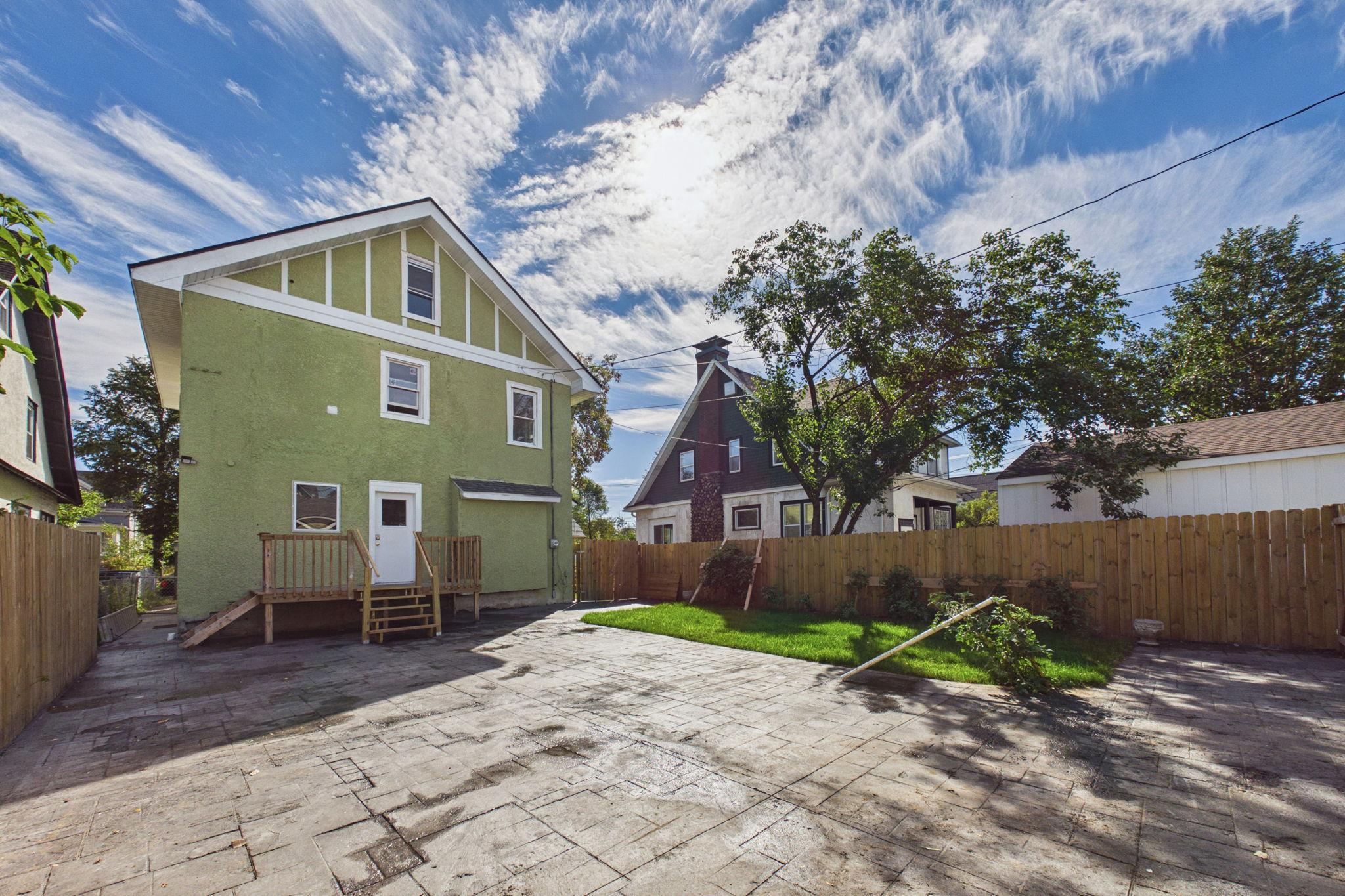 Green house viewed from a paved backyard with a small lawn and wooden privacy fence.