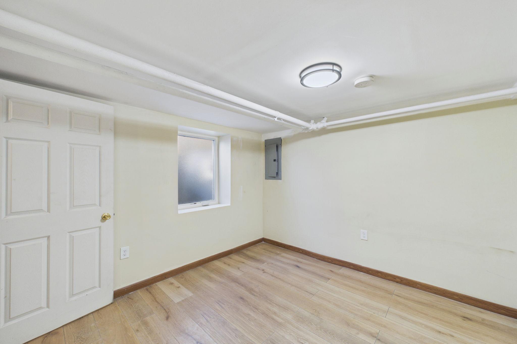 Empty room with light plank flooring, a small window, and exposed ceiling pipes.