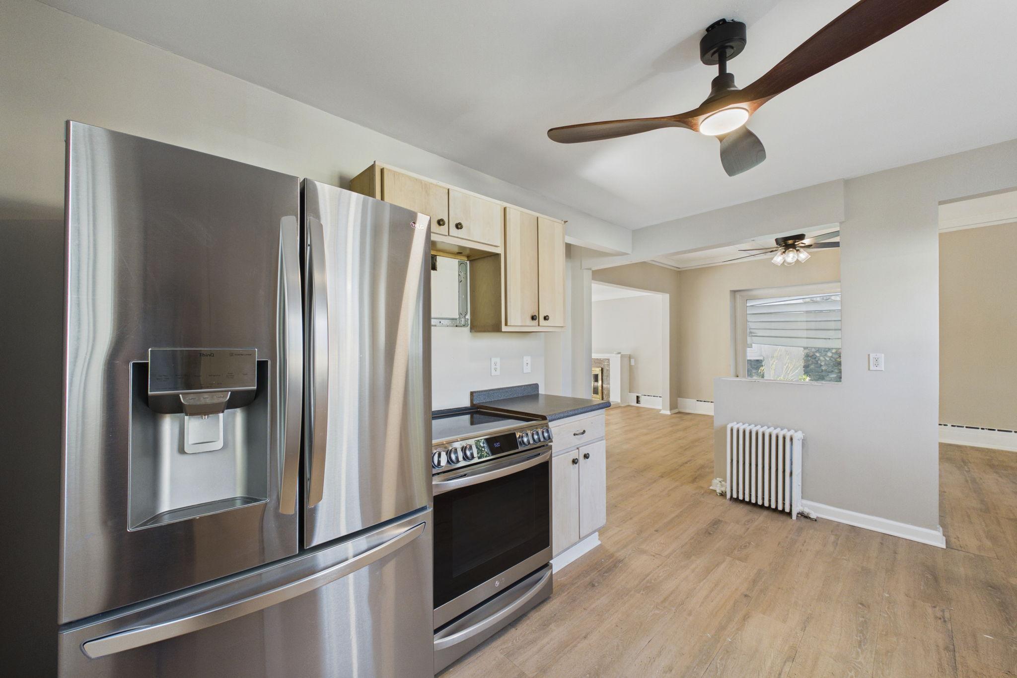 Kitchen with stainless steel appliances, pale wood cabinets, and flooring that continues into an open adjoining room.