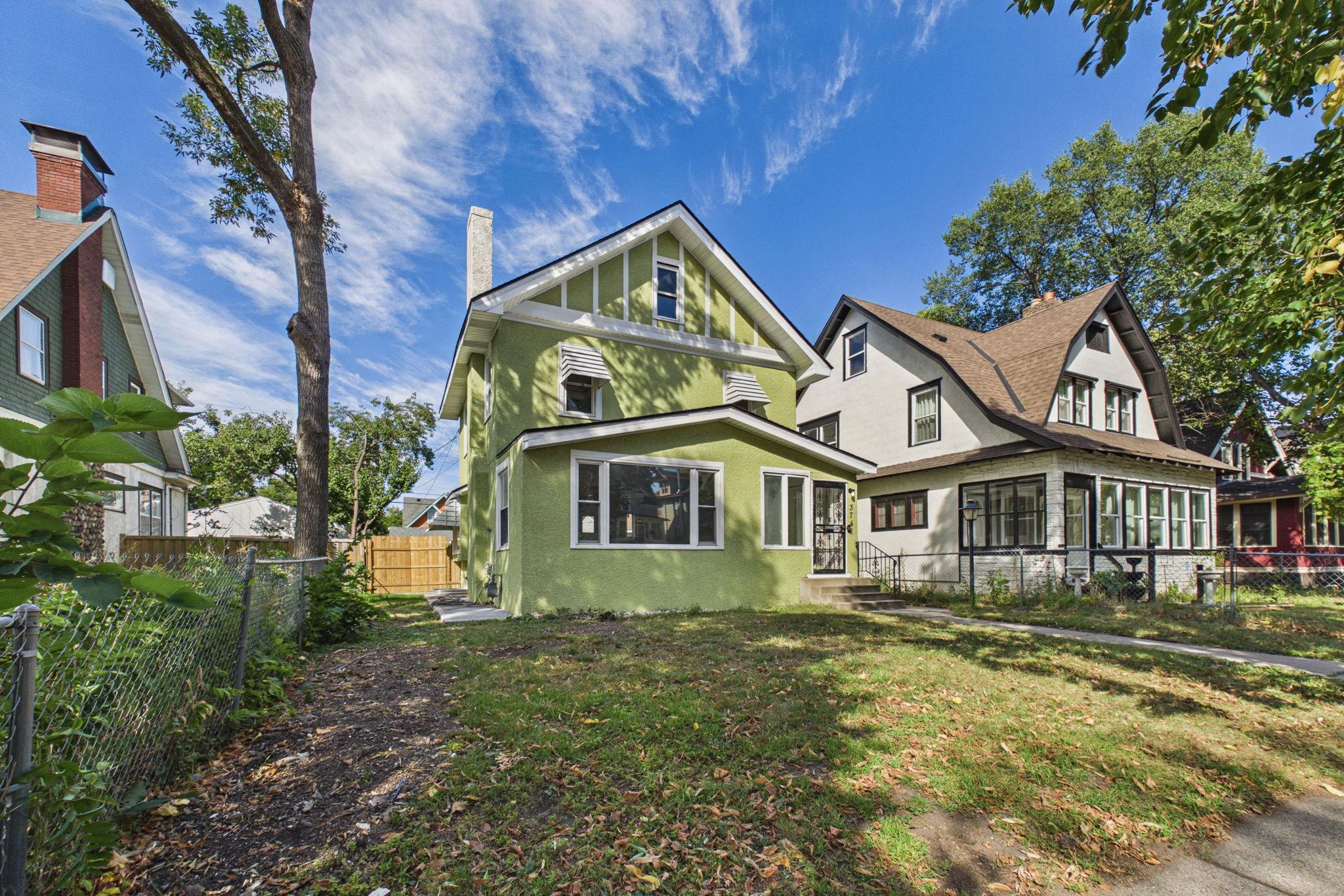 Green two-story house with white trim and a steep gabled roof, viewed from its fenced side yard.