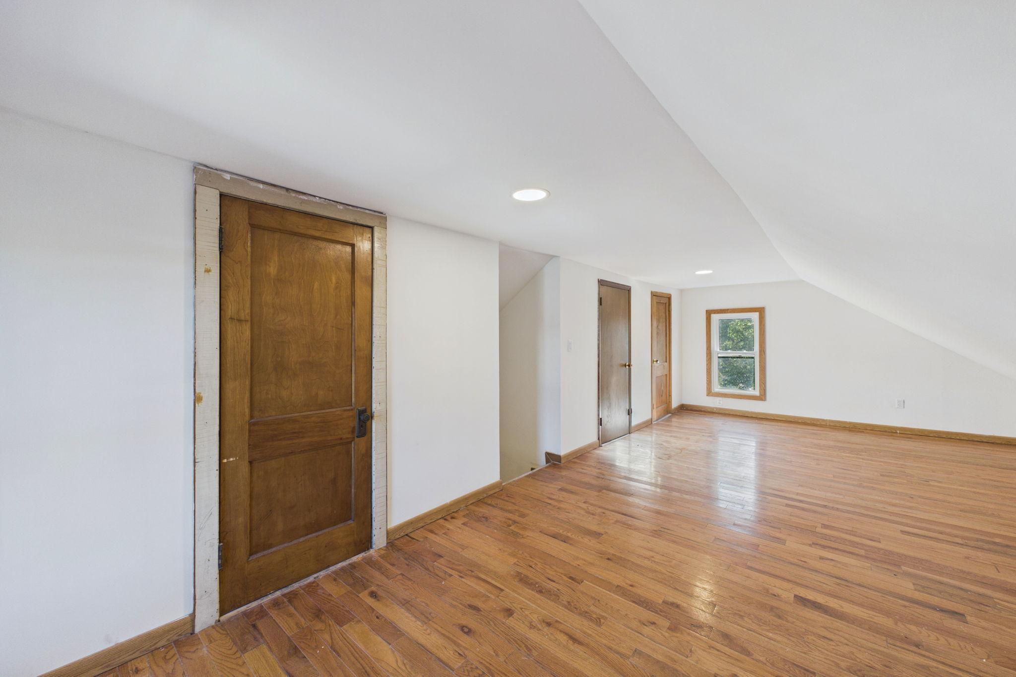 Empty upper-floor room with hardwood floors, a sloped ceiling, and several wooden doors.